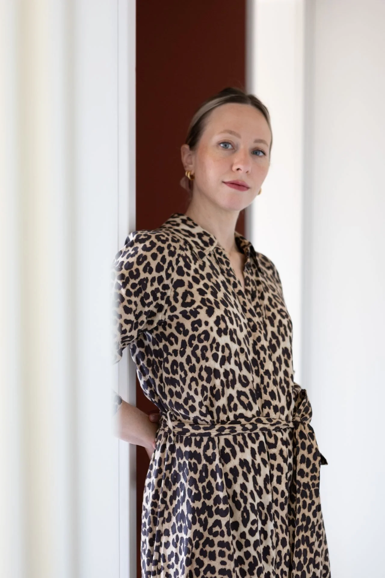 A woman with blue eyes and blonde hair, wearing a leopard print dress and gold hoop earrings, standing by a doorway with a neutral expression.