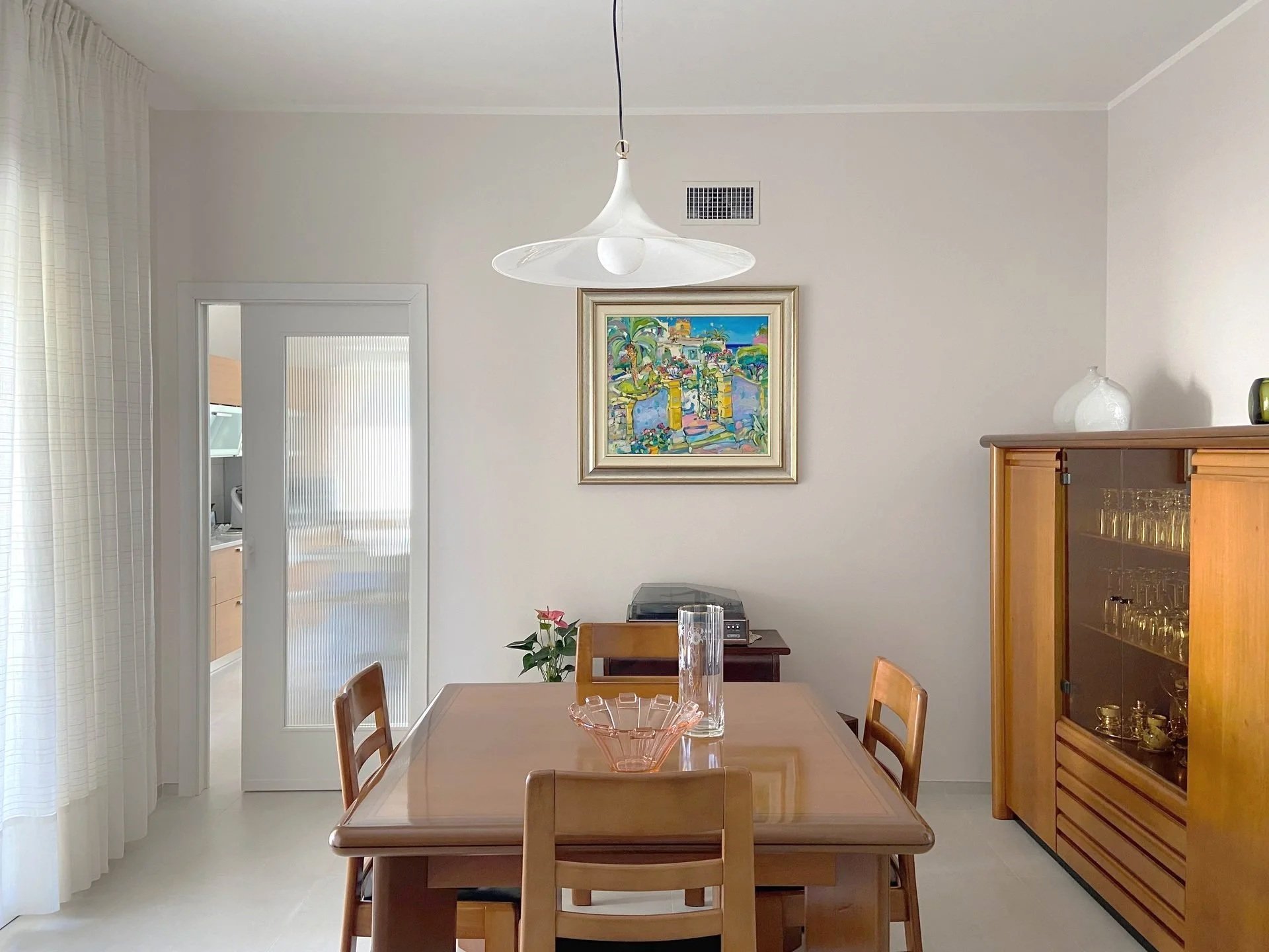 A dining room with a wooden table and chairs, a glass and pink bowl on the table, a potted plant, artwork on the wall, and a wooden cabinet with glass doors. There is a door with frosted glass and curtains on the left and various vases on top of the cabinet.