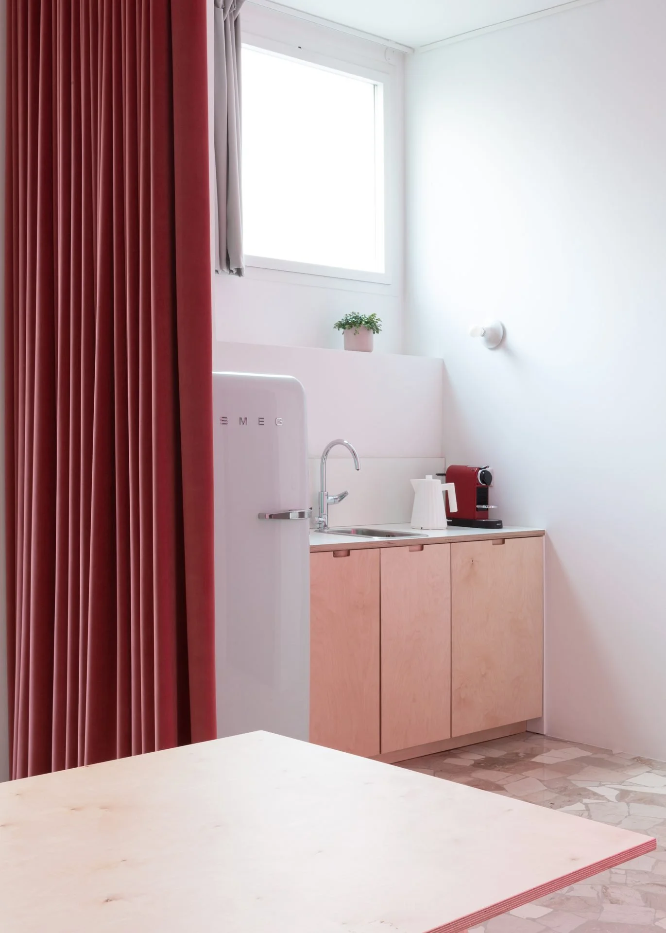 A kitchen with a white countertop, light pink cabinets, a small potted plant on the window sill, a red coffee machine, a white electric kettle, and red curtains covering a window.