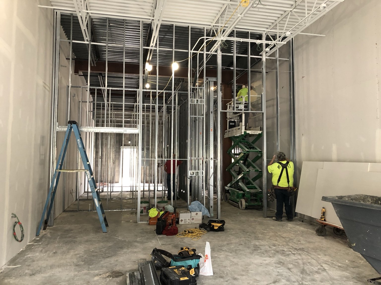 Construction workers are building the steel framing of a room, with tools and equipment on the unfinished floor, including a blue ladder on the left and a scissor lift on the right.