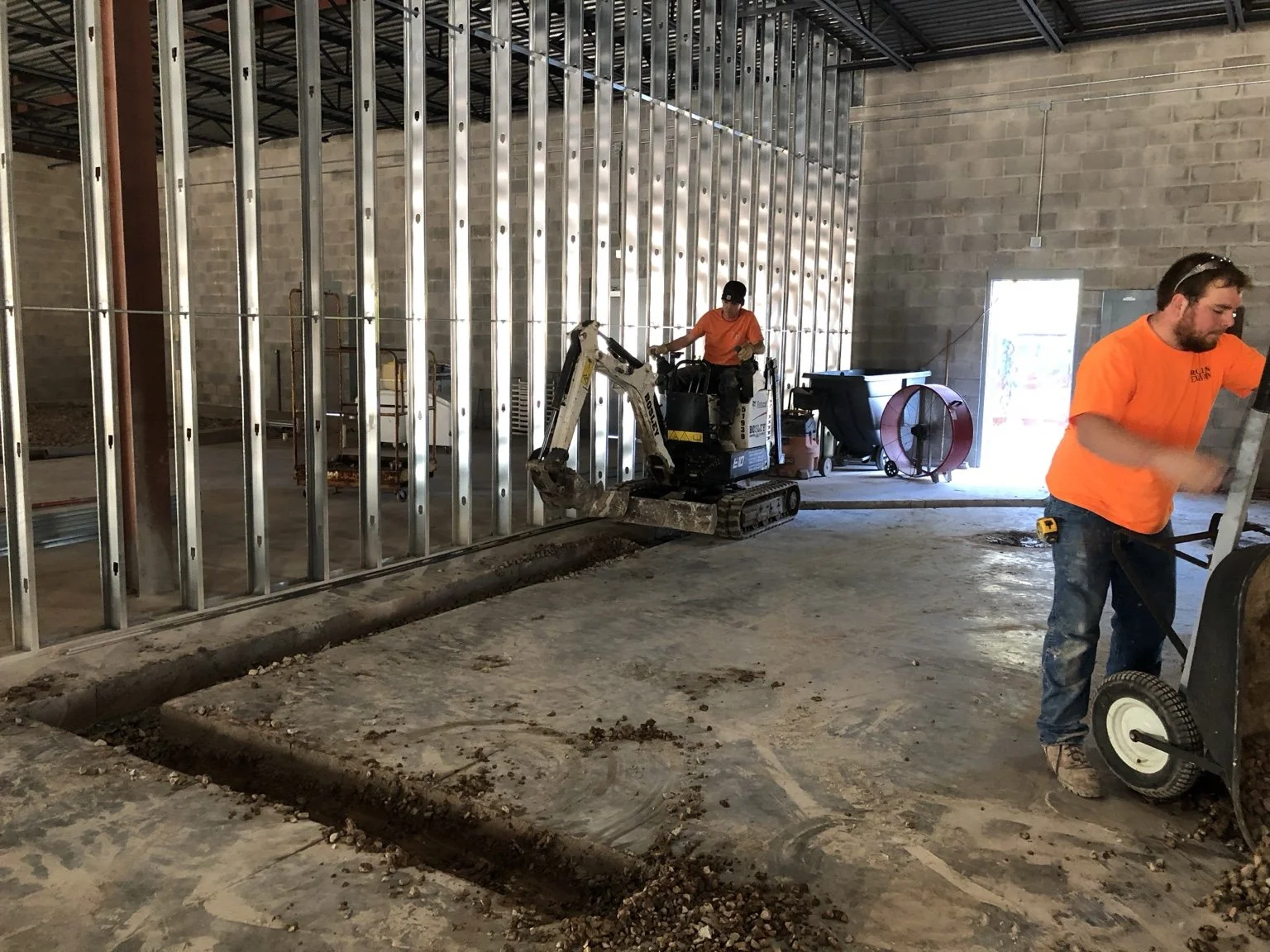 Construction workers installing metal wall studs inside a building. One worker operates a mini excavator, while another uses a wheelbarrow.