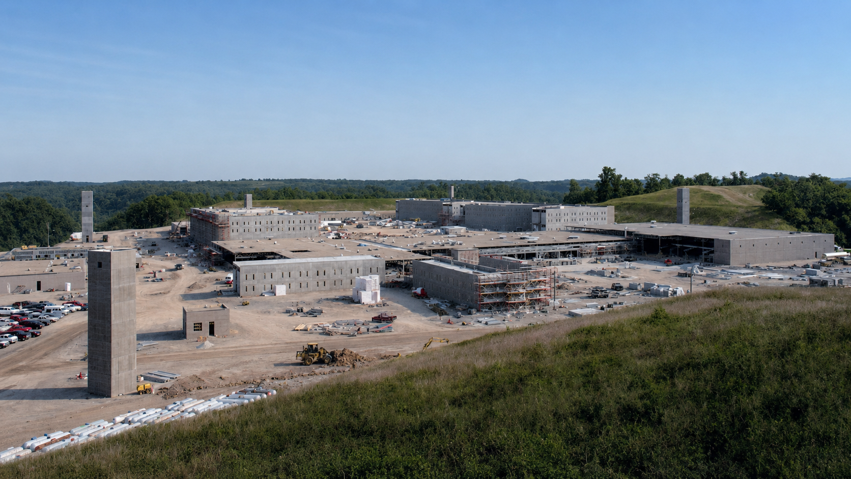 A large construction site with multiple buildings in progress, surrounded by trees and hills, with construction vehicles and parking areas visible in the foreground.