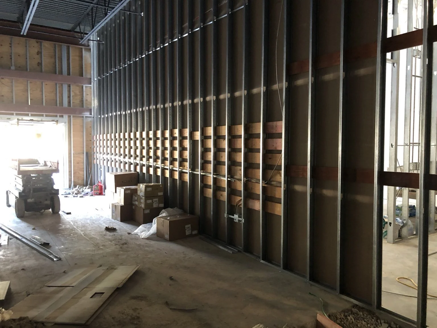 Interior of a building under construction, showing exposed metal wall studs, electrical wiring, and construction materials and boxes on the floor.