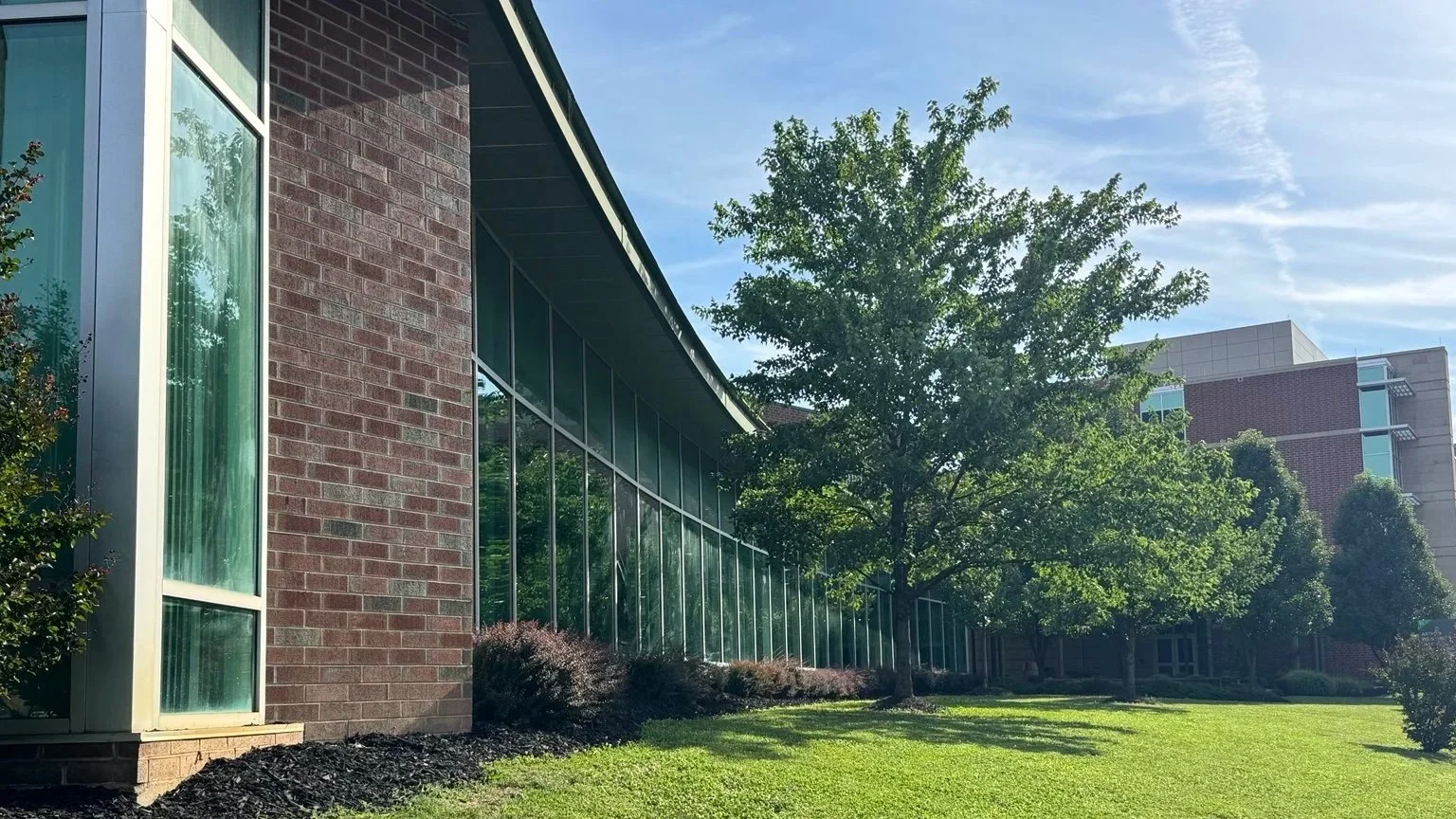 Exterior view of the Arthur Weisberg Family Applied Engineering Laboratories at Marshall University with large glass windows, a brick wall, and greenery including grass and trees, under a partly cloudy sky.