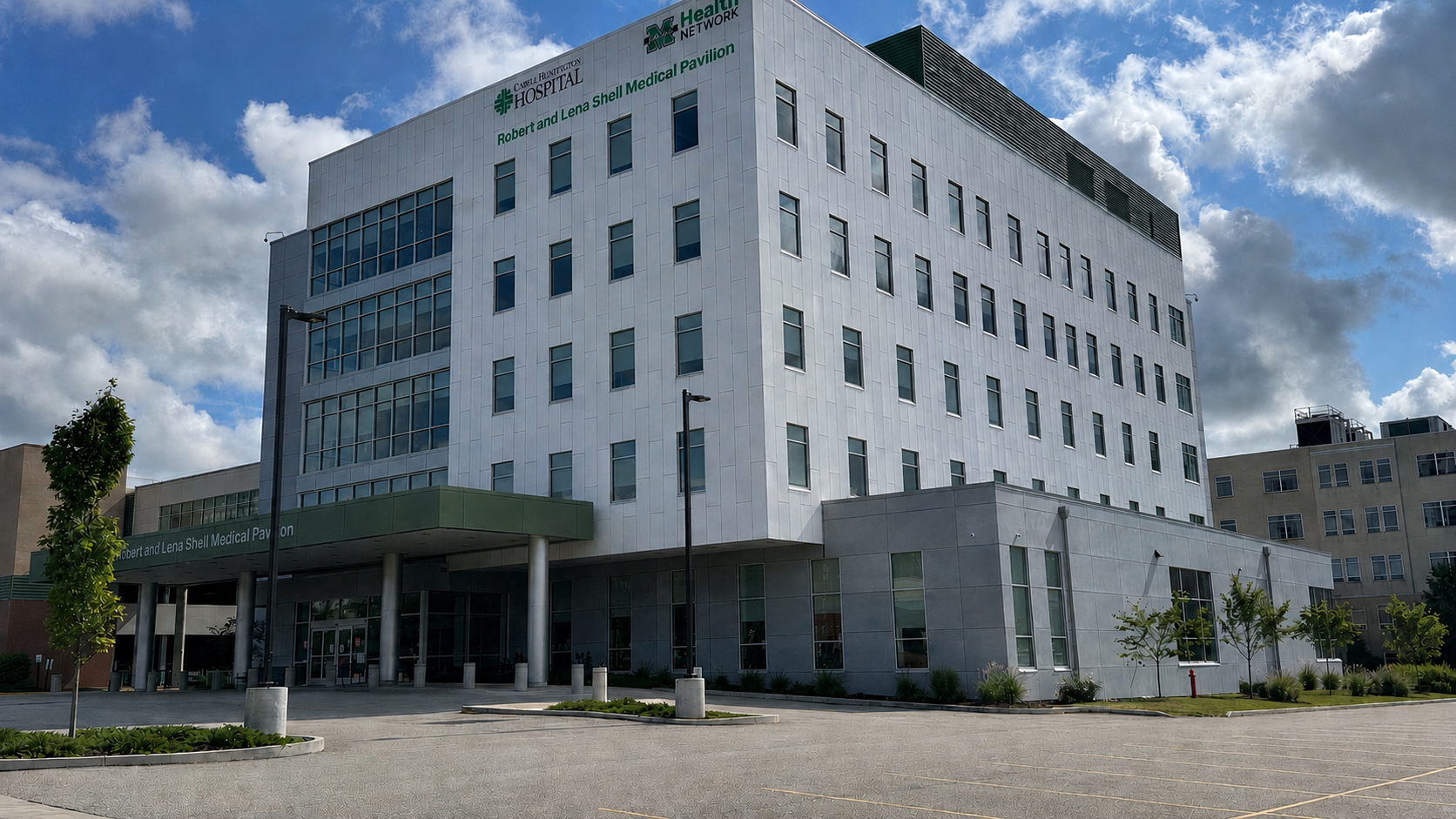 Exterior view of the Robert and Lena Shell Medical Pavilion, a modern multi-story hospital building with large windows and a white facade, surrounded by a paved driveway, trees, and street lamps under a partly cloudy sky.