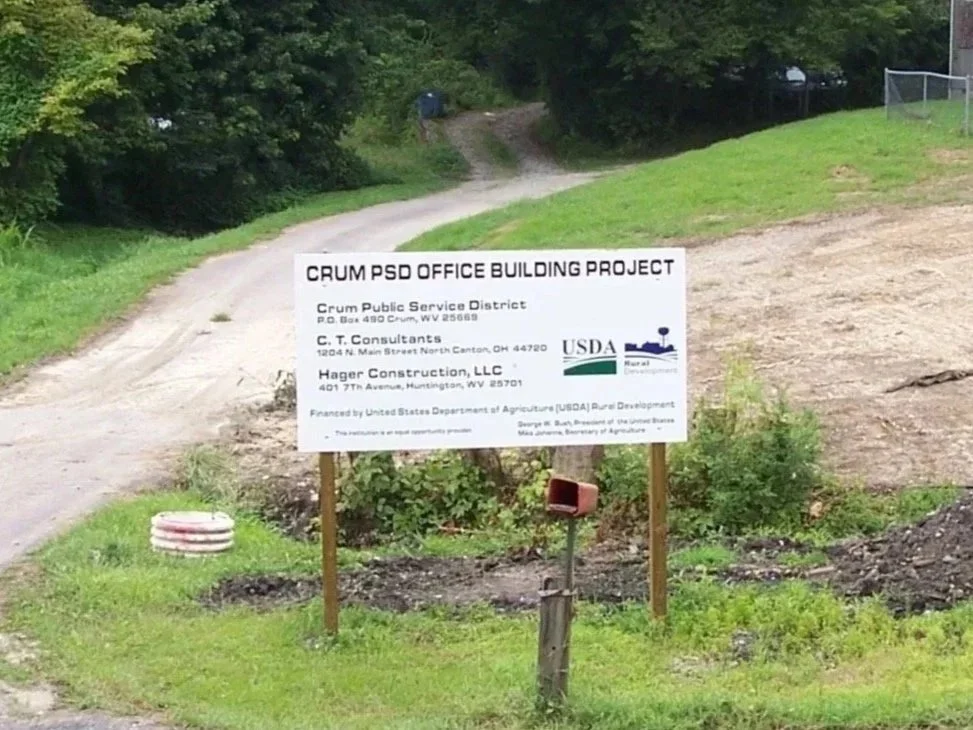 Signboard at a construction site with information about the Crum PSD Office Building Project, including addresses and contractors, surrounded by green grass and trees, with a dirt road in the background.
