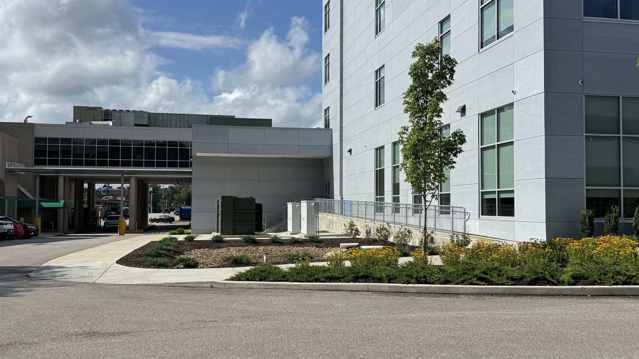 Exterior of Robert and Lena Shell Medical Pavilion with large windows, a small tree, and a landscaped area with yellow flowers in the foreground. Parking lot and cars visible beneath elevated walkway.