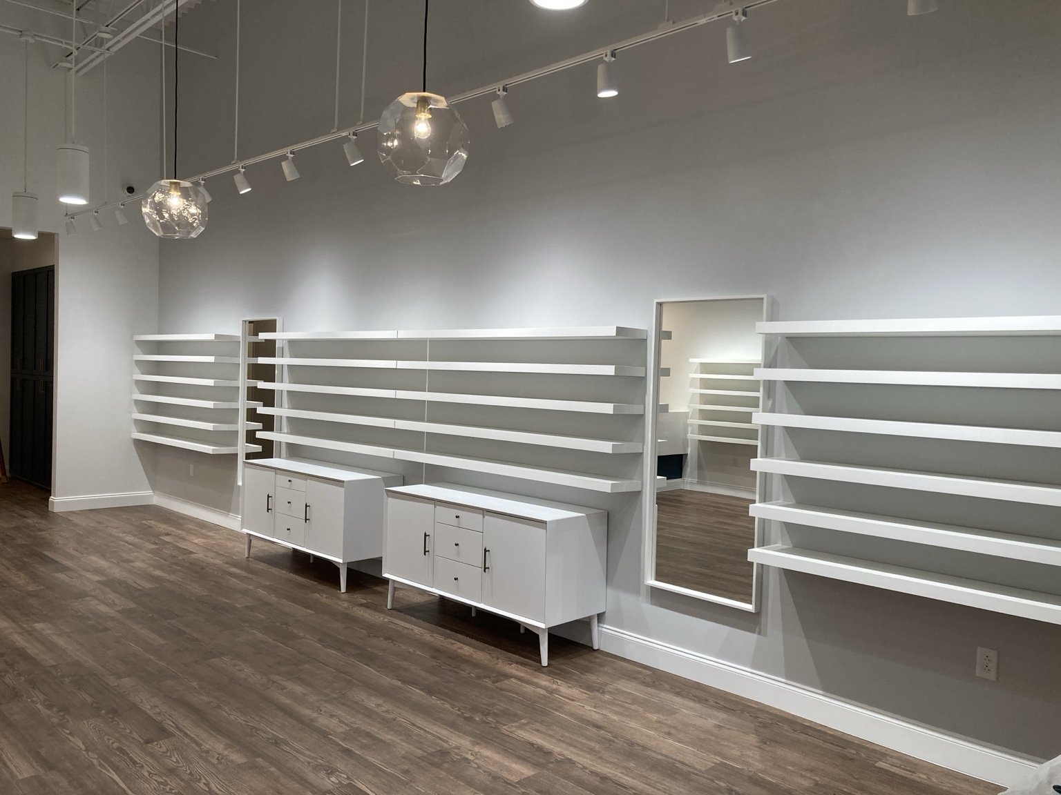 Empty interior space with white shelves, a large mirror, wooden flooring, and modern lighting fixtures.
