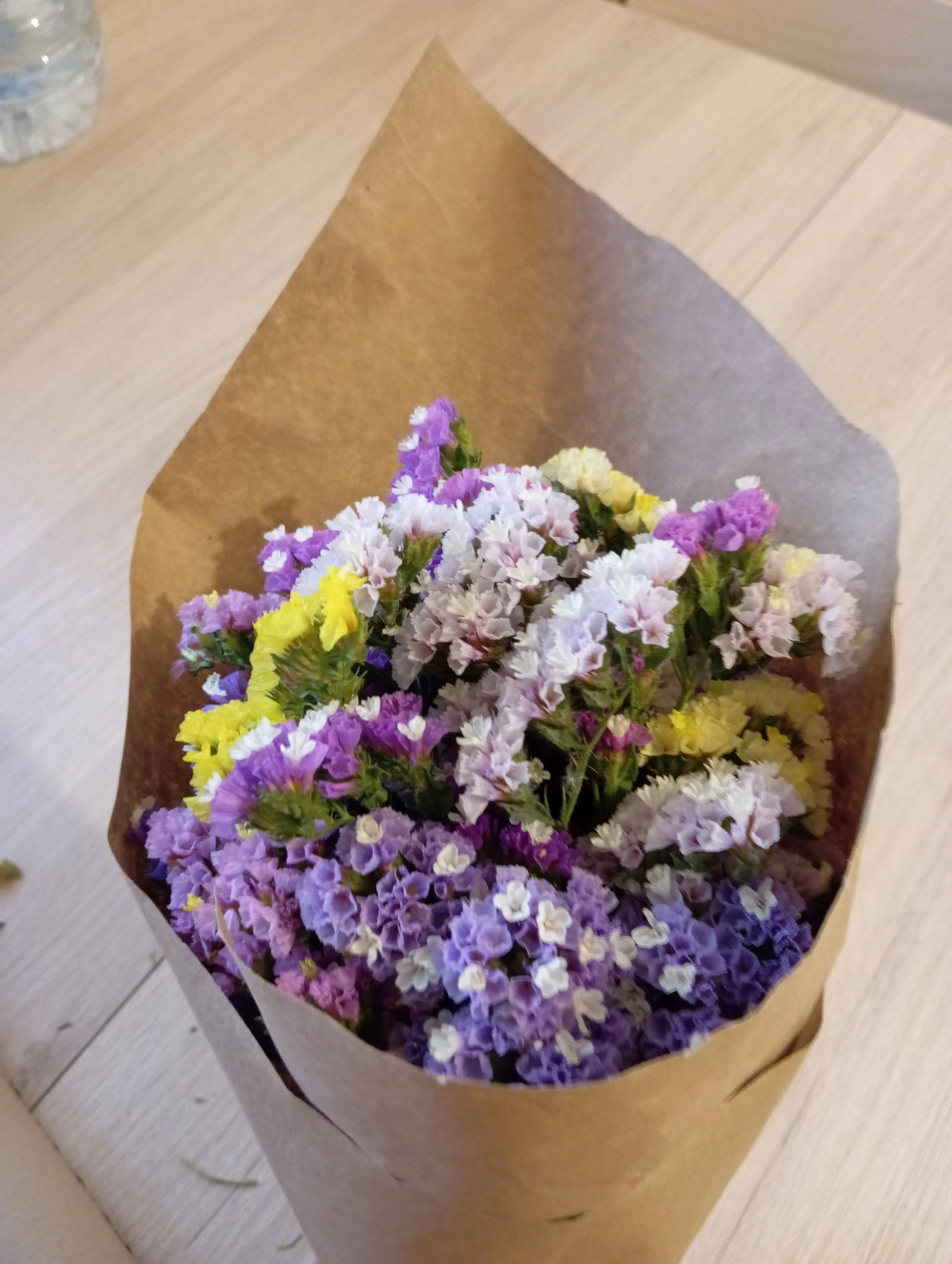 Ramo de flores de colores rosados, blancos, morados y amarillos envuelto en papel kraft, colocado sobre una mesa de madera.
