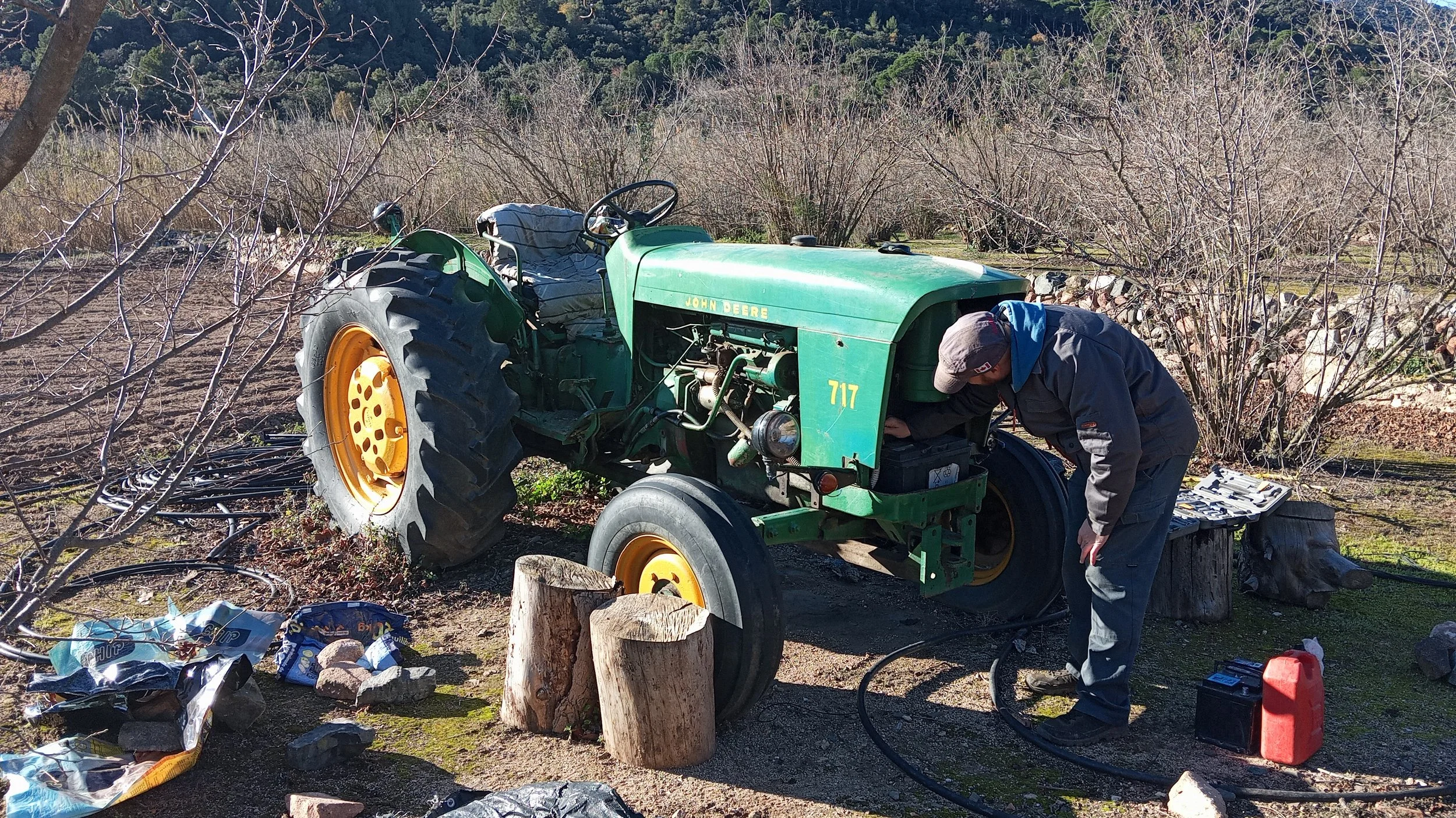 Una persona arreglando un tractor John Deere verde en un campo con arbustos y árboles sin hojas, junto a herramientas y objetos esparcidos en el suelo.