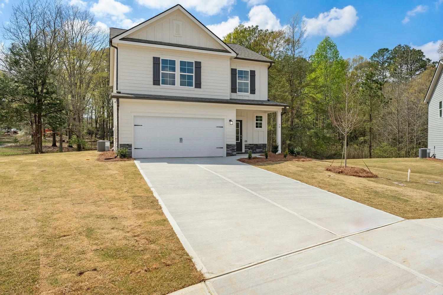 New two-story house with a white garage door, gray roof, front porch, and a small yard with a young tree, situated in a neighborhood with similar houses, under a blue sky with clouds.