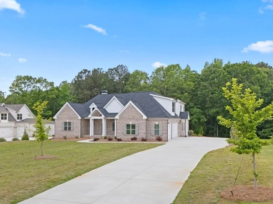 A two-story house with brick and white siding exterior, a front porch, and a curved concrete driveway, surrounded by a lawn with newly planted trees, with forested trees in the background and a blue partly cloudy sky.