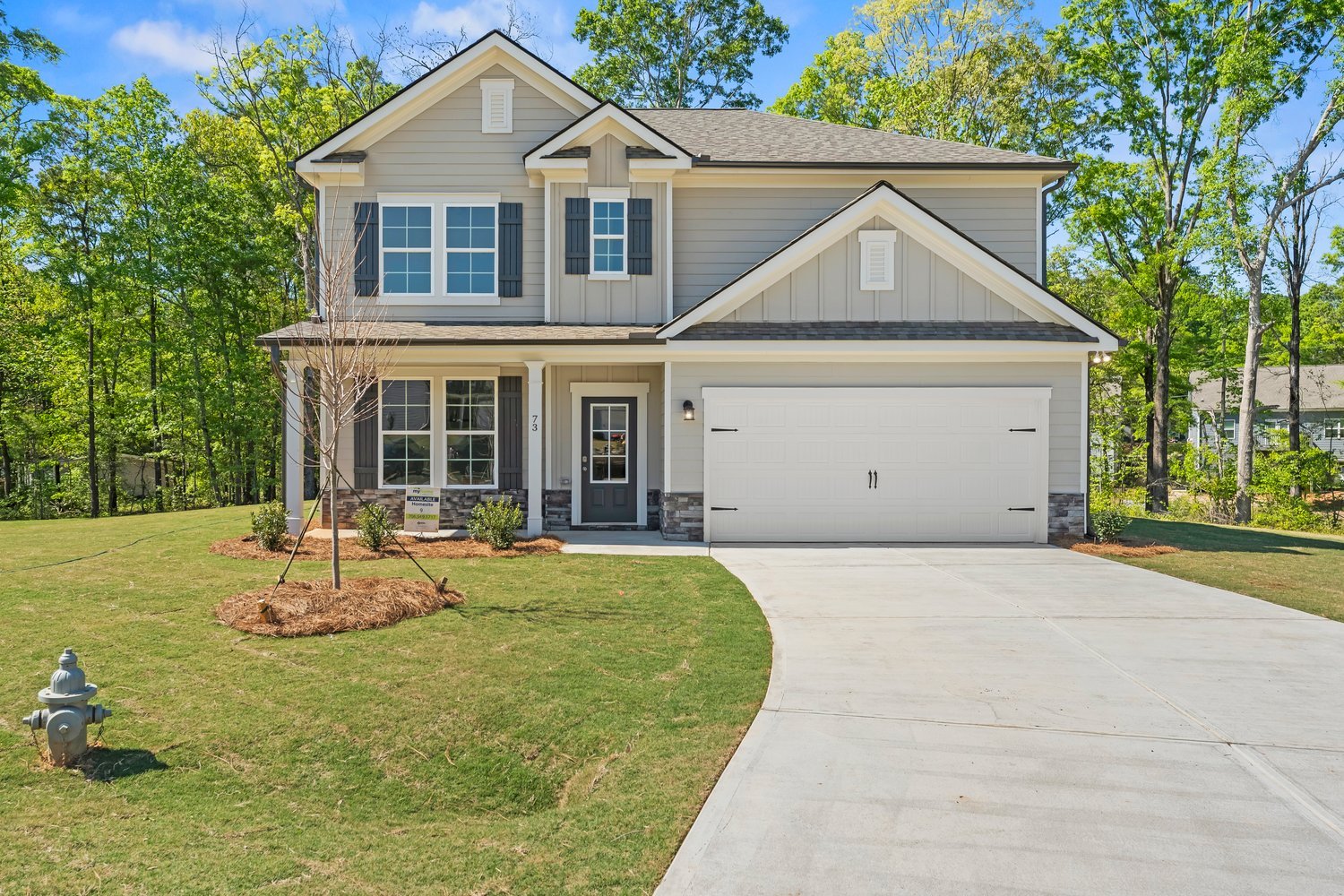 New two-story house with gray siding, white trim, black shutters, and a two-car garage. Front yard with young trees and landscaping, concrete driveway, and surrounded by green trees.