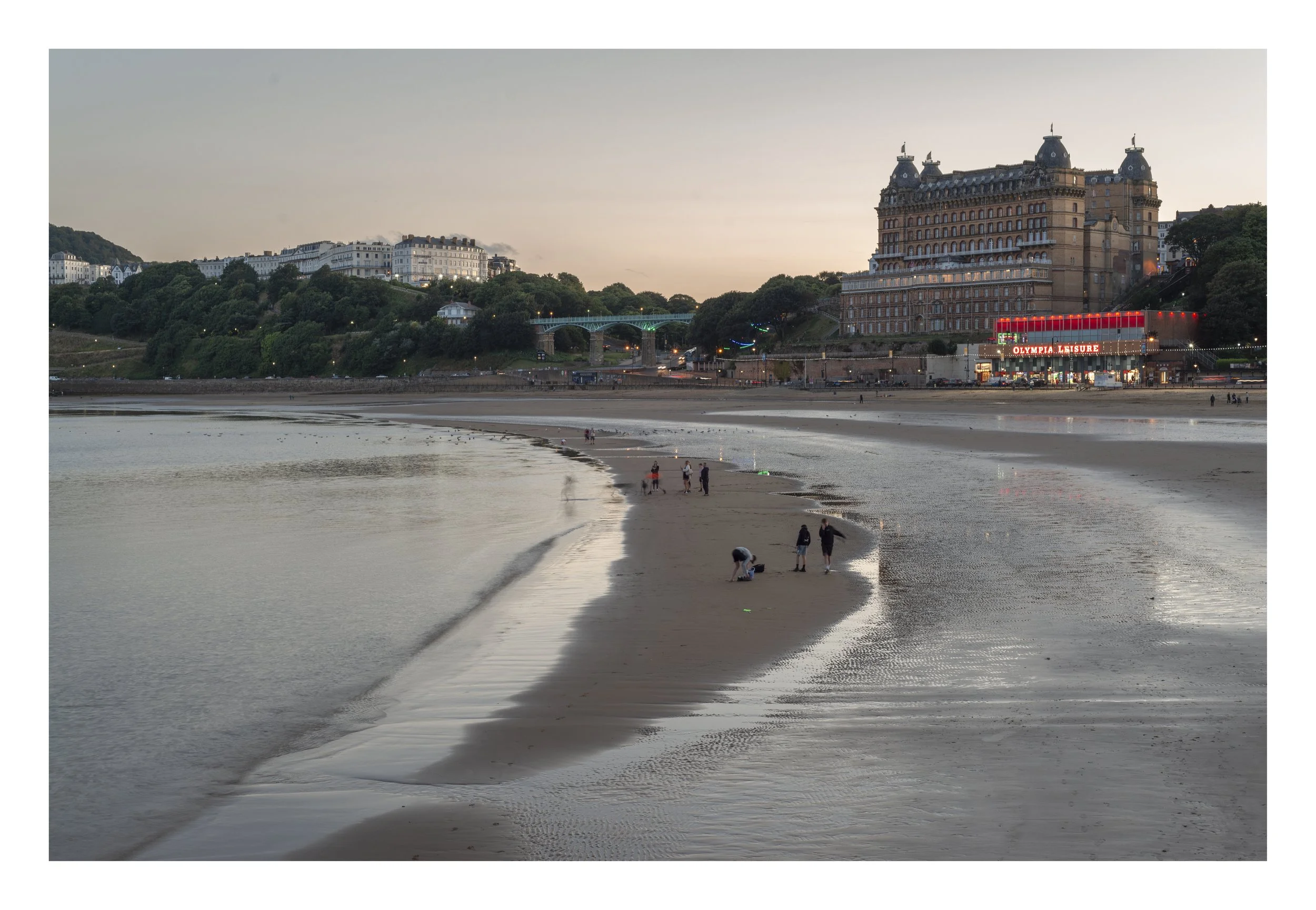 A beach scene at sunset with people walking and relaxing, a historic hotel with bright neon lights reading 'Olympia Leisure,' a bridge over green hills, and buildings in the background.