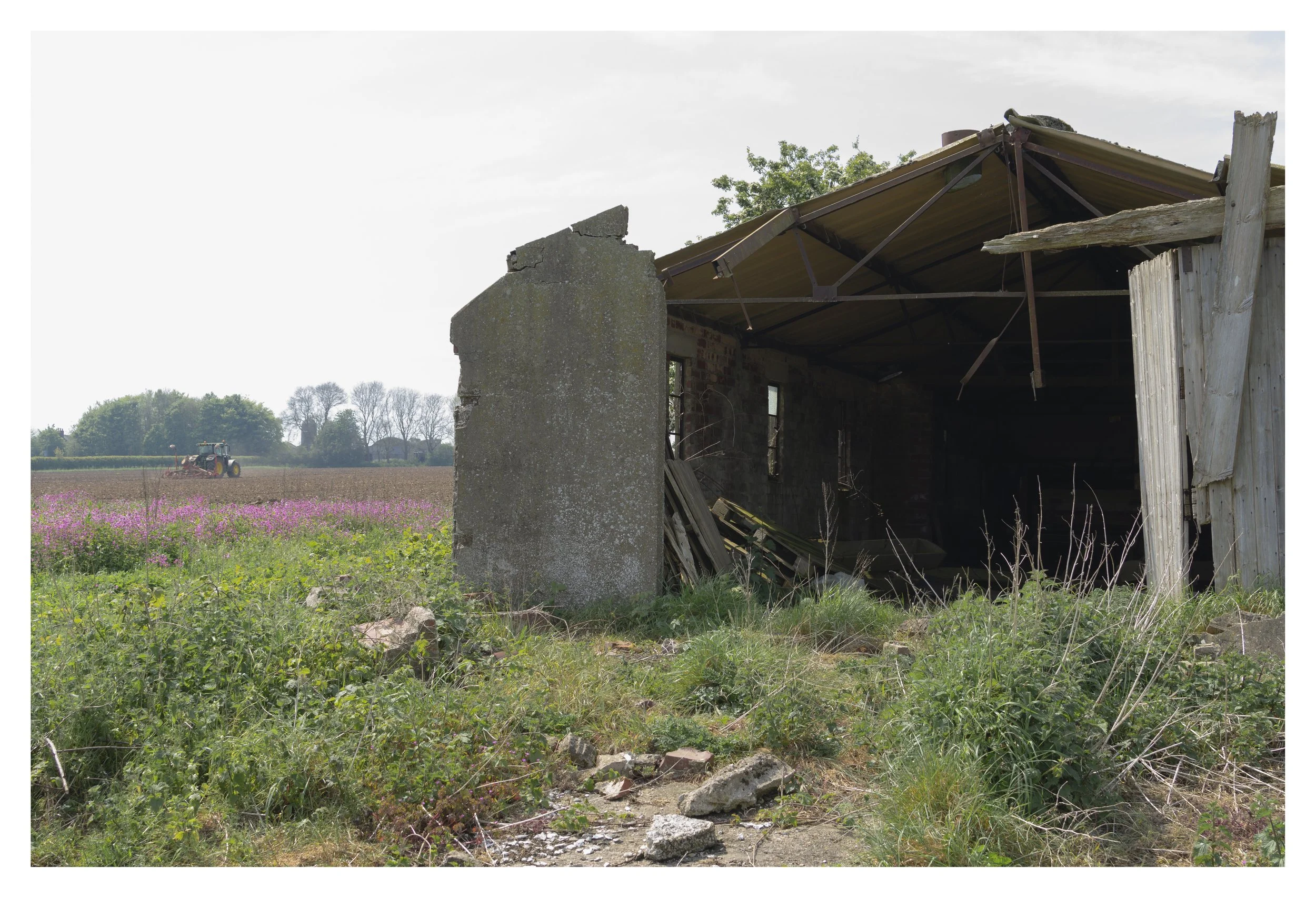 An abandoned building with a partially collapsed roof, surrounded by overgrown grass and weeds, with a field and tractor in the background under a cloudy sky.