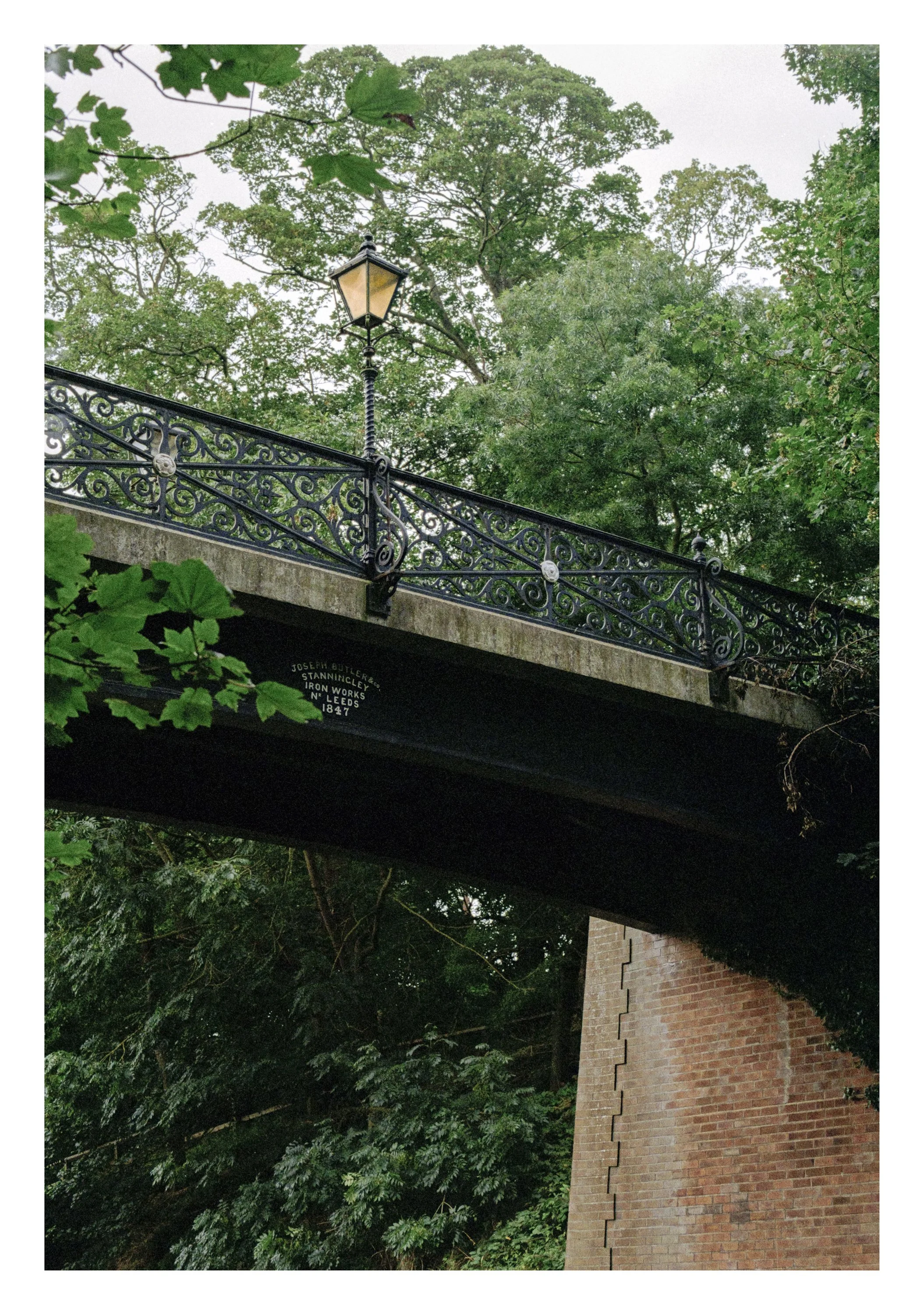 A street view showing a metal bridge with ornate black iron railings and a street lamp, surrounded by green trees and a brick wall beneath the bridge.