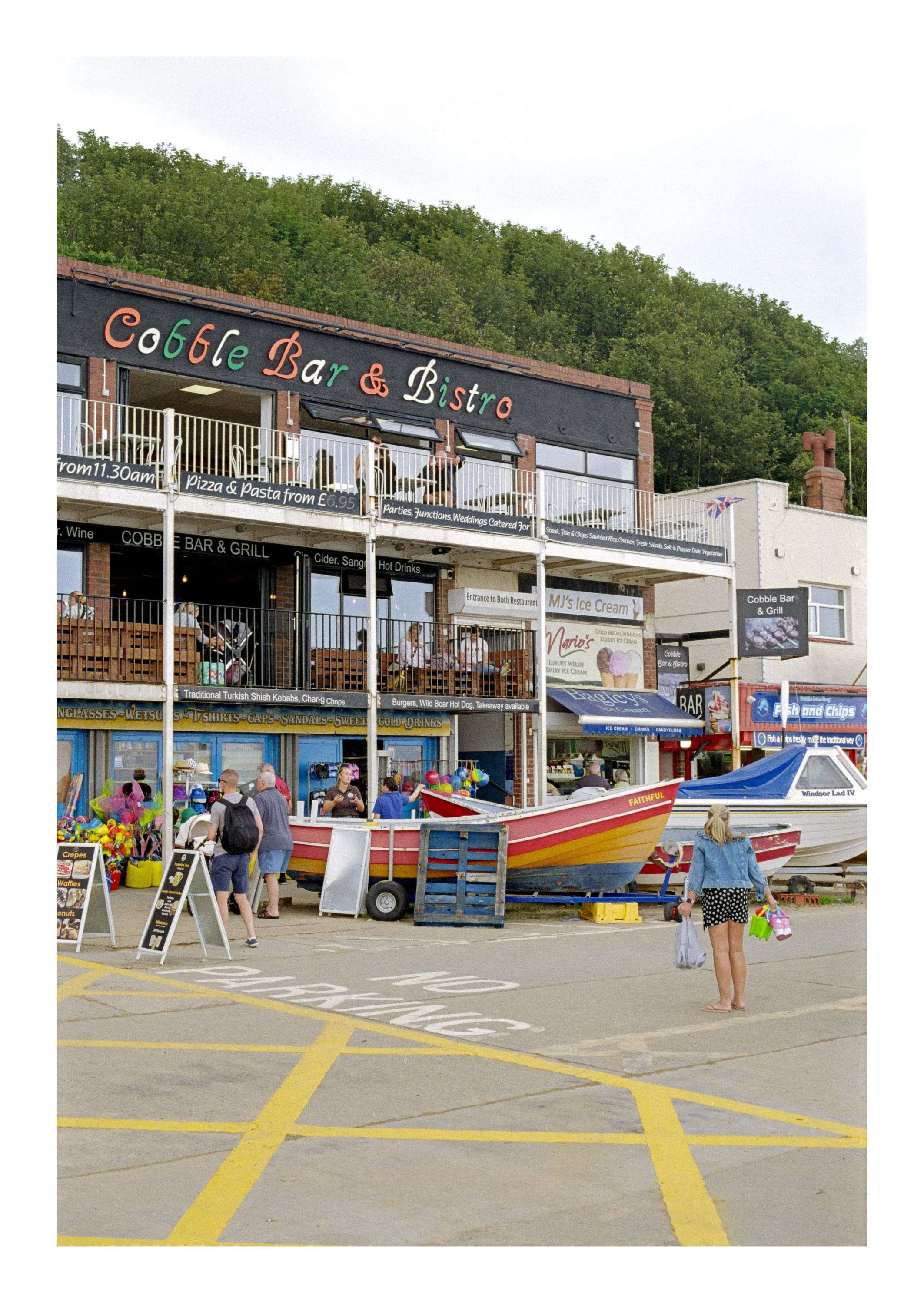 People walking in a parking lot near boats, with a building labeled 'Cobble Bar & Bistro' on the upper level, and various signs advertising food and drinks. A boat named 'FAITHFUL' is in the foreground.