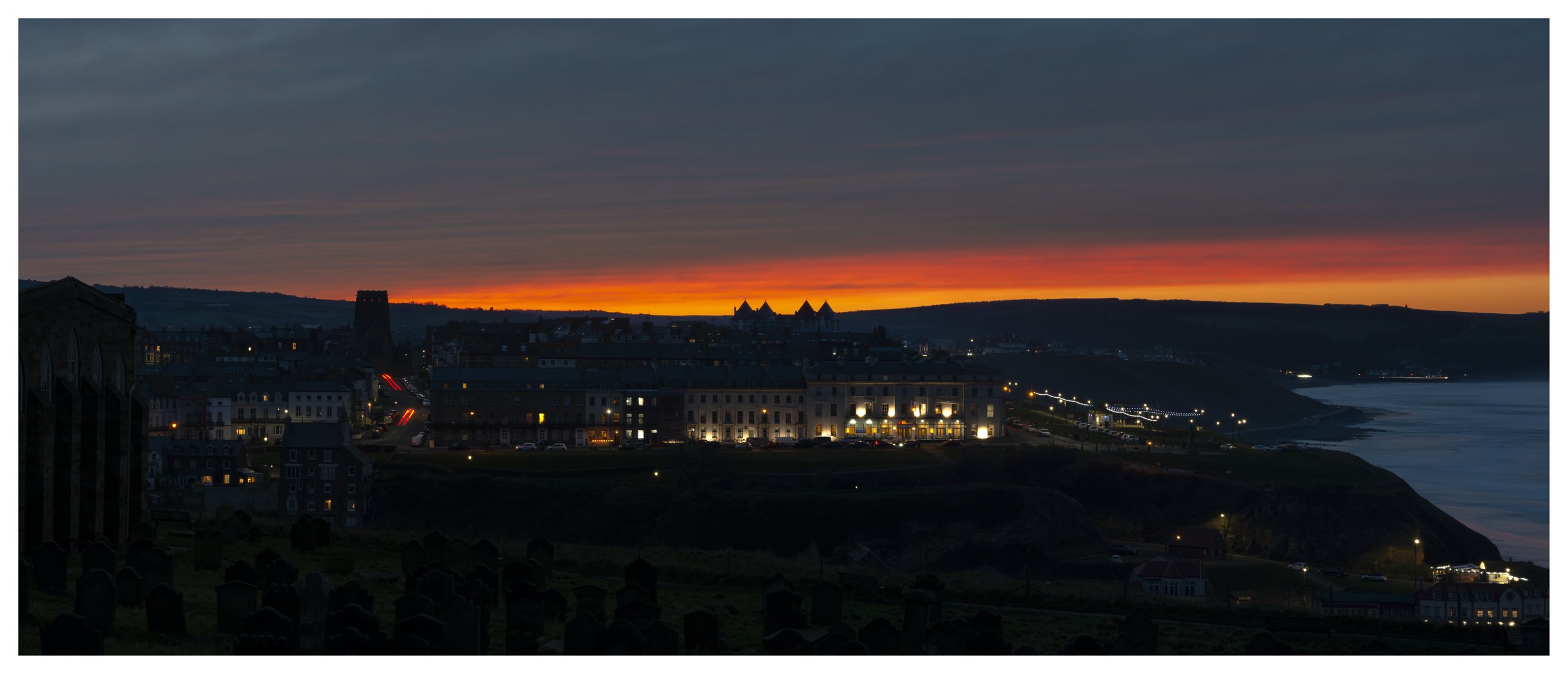 Sunset over a town with illuminated buildings along the coast, dark clouds in the sky, and streaks of red and orange near the horizon.