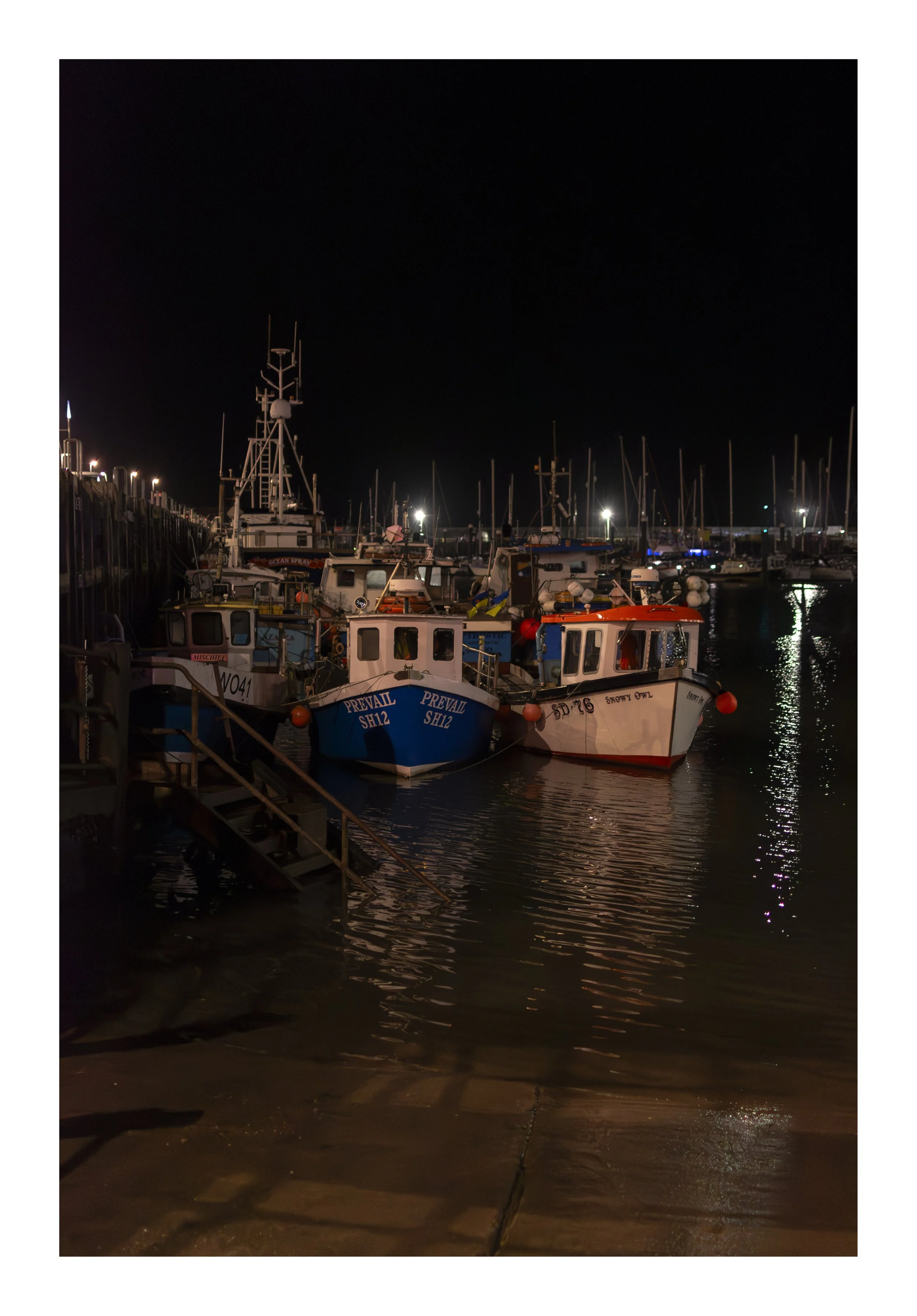 Nighttime scene at a marina with boats docked along the pier, illuminated by small lights, with reflections on the water, and a dark sky overhead.