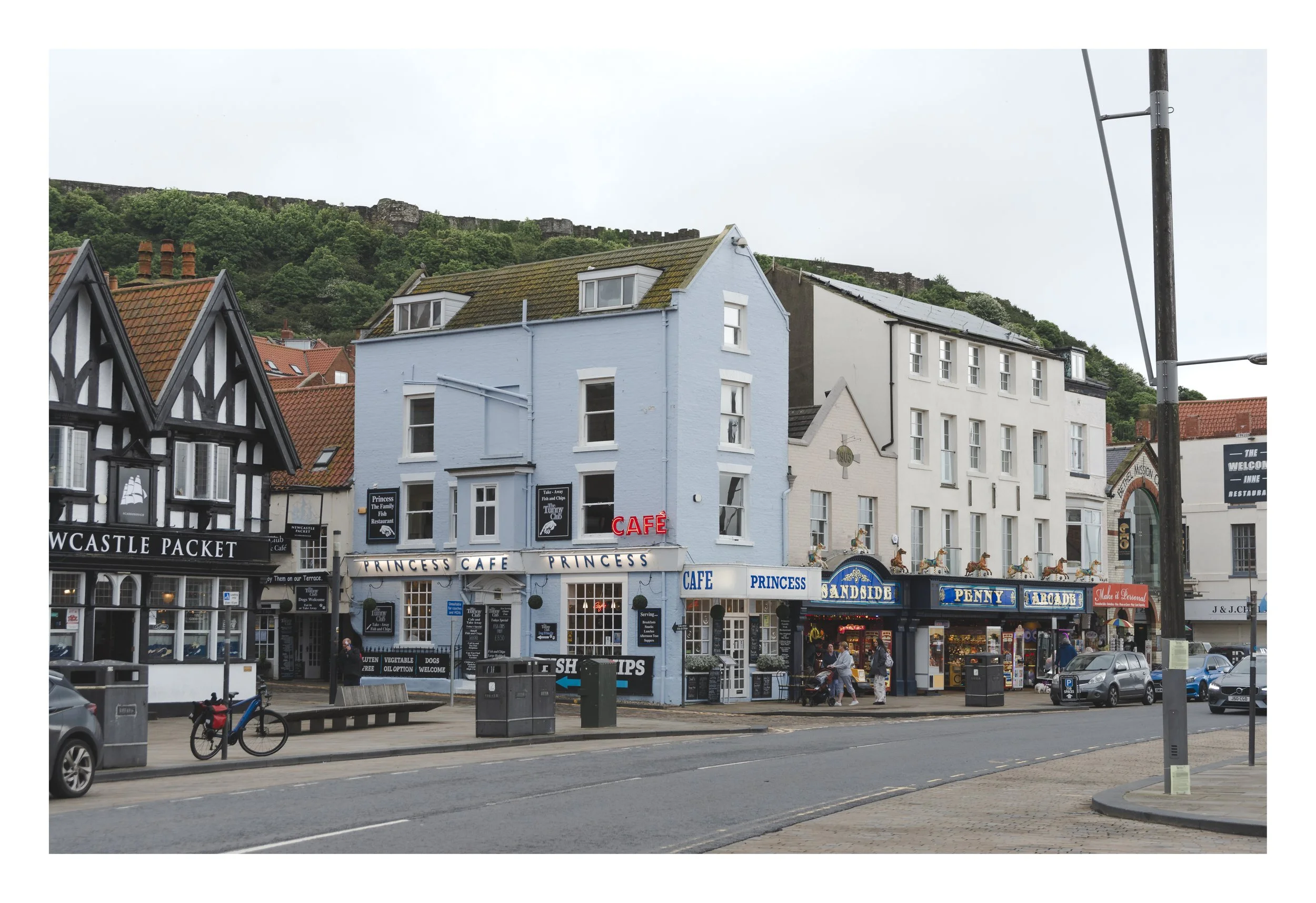 Street view of shops and cafes with a hilltop fortress in the background, overcast sky.