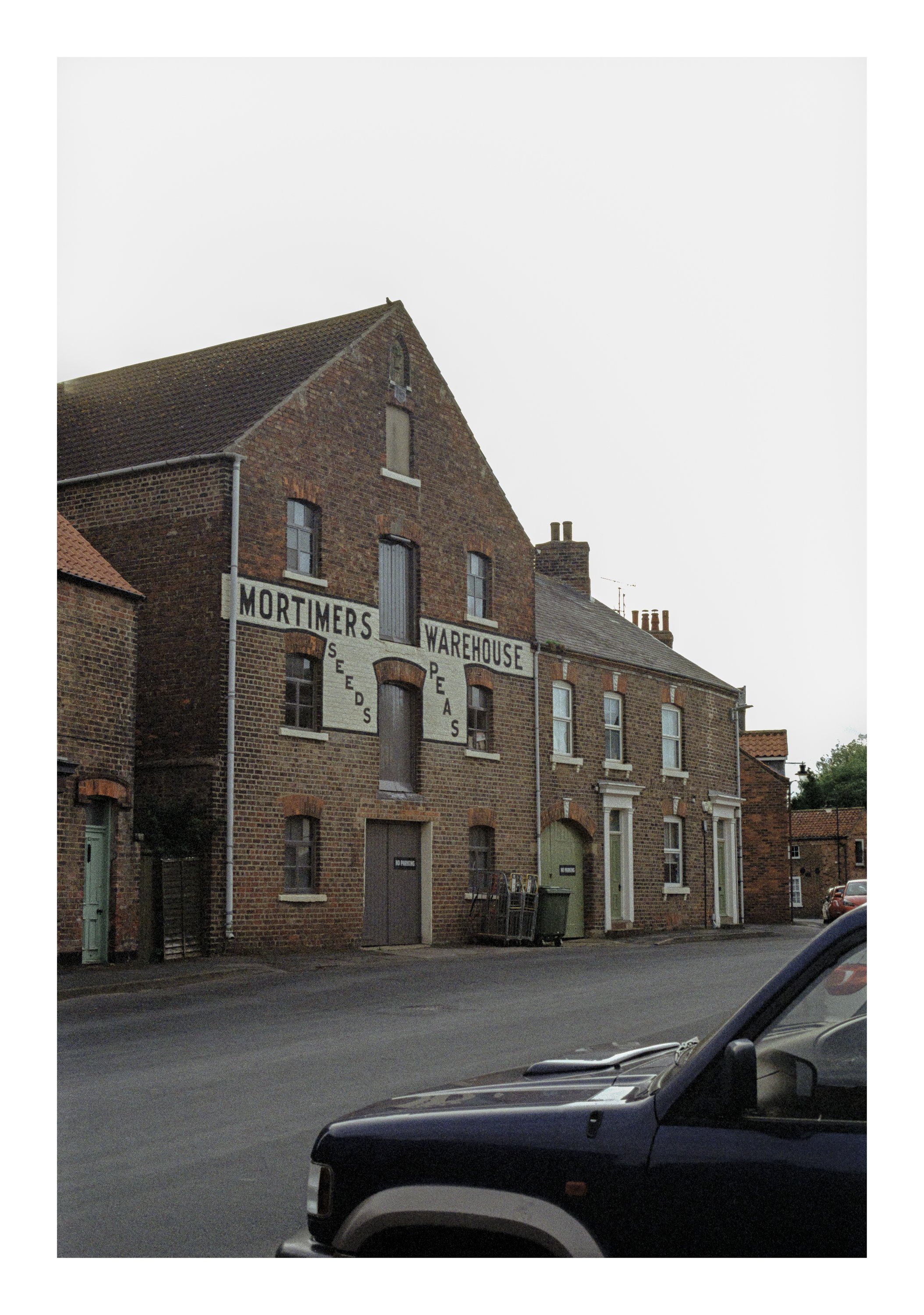 A street view showing a brick building with a faded sign that reads 'Mortimers Warehouse'. The building has multiple windows, and there are some trash bins on the sidewalk. Part of a parked car is visible in the lower right corner.