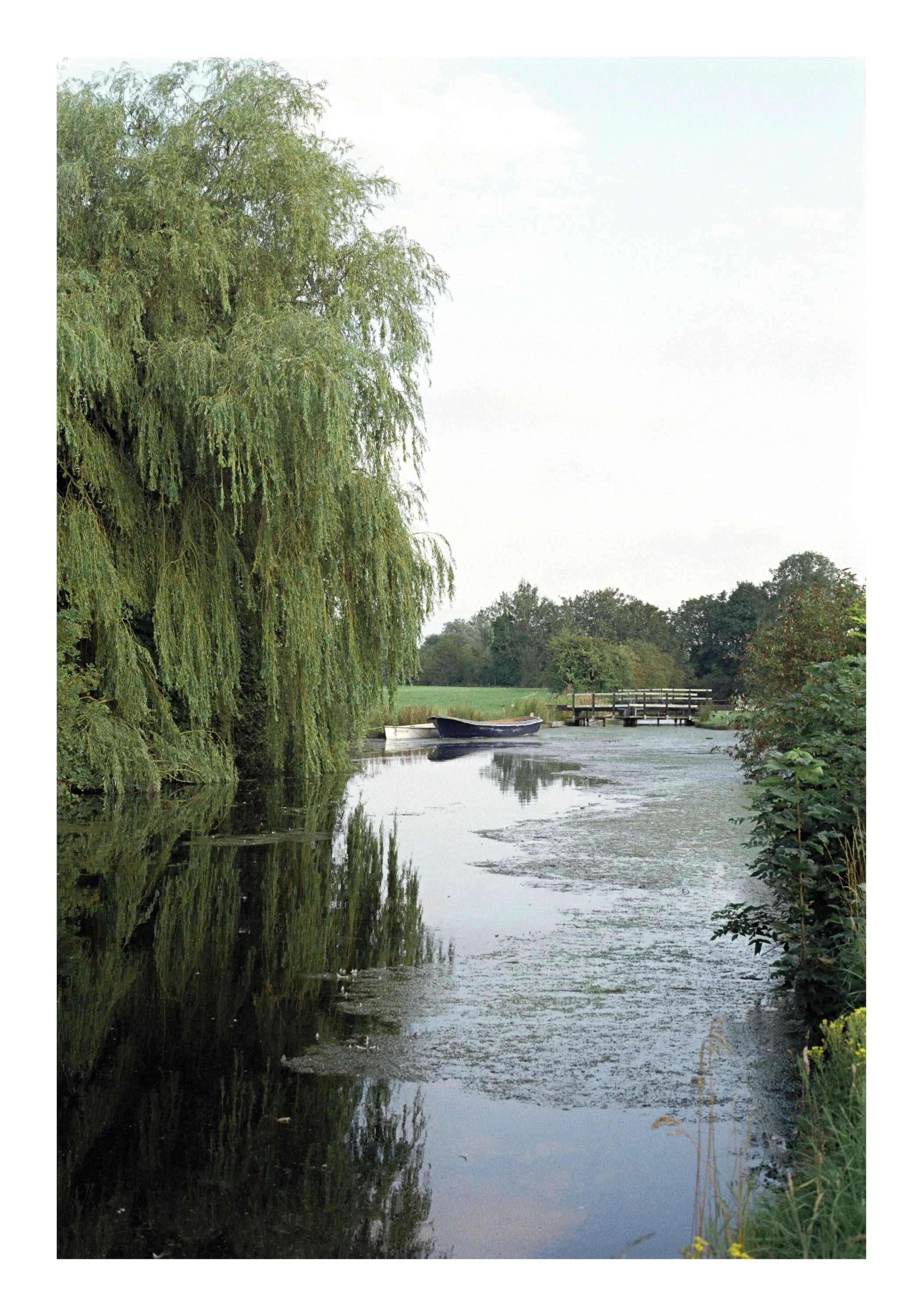 A peaceful scene of a canal with calm water reflecting the trees along the banks, a small boat docked at the edge, and a wooden pier in the background, surrounded by lush greenery under a partly cloudy sky.