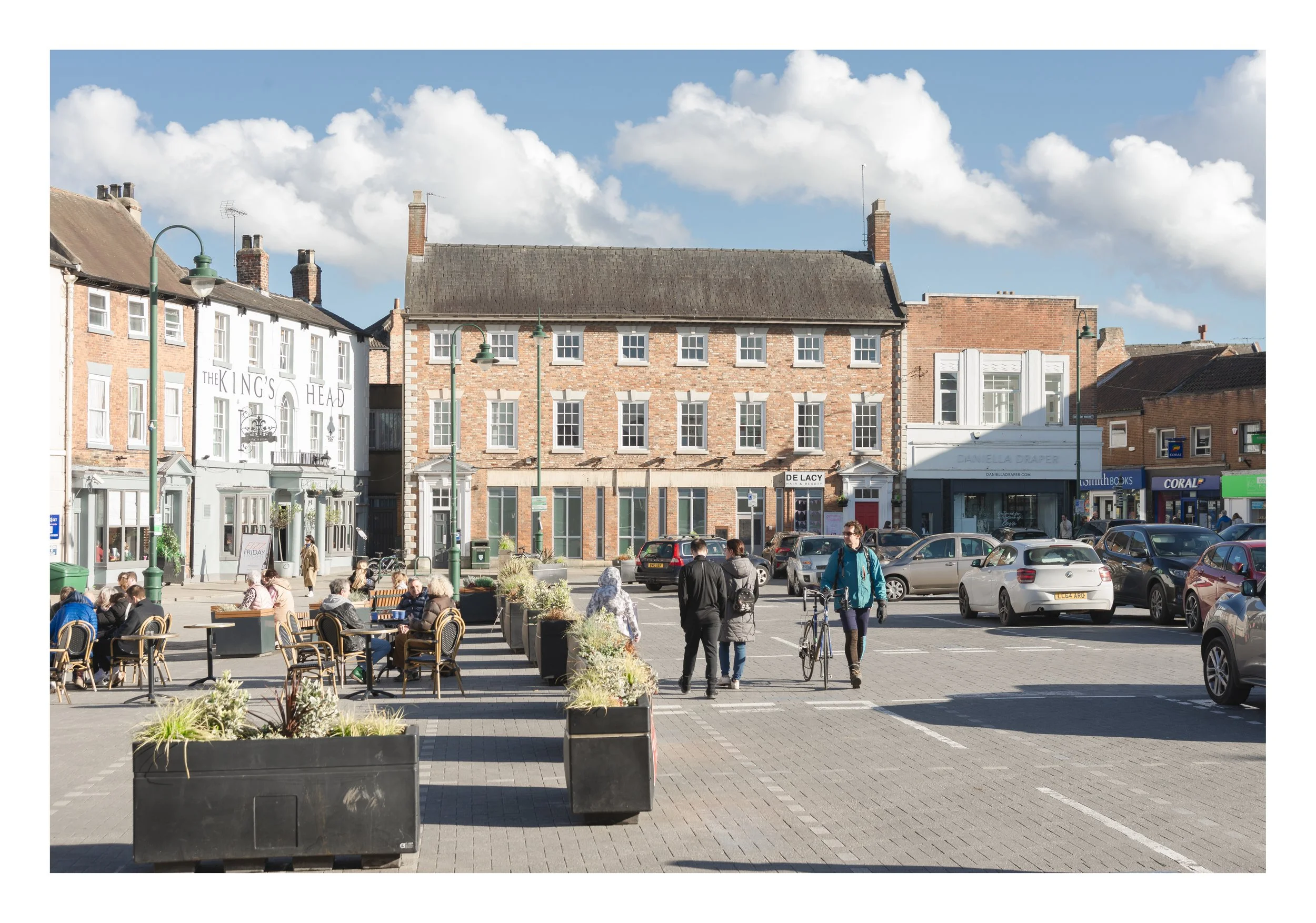 A lively street scene in daytime featuring outdoor dining with groups of people seated at tables, parked cars lining the street, and brick buildings with a mix of white and brick facades. The sky is blue with scattered clouds.