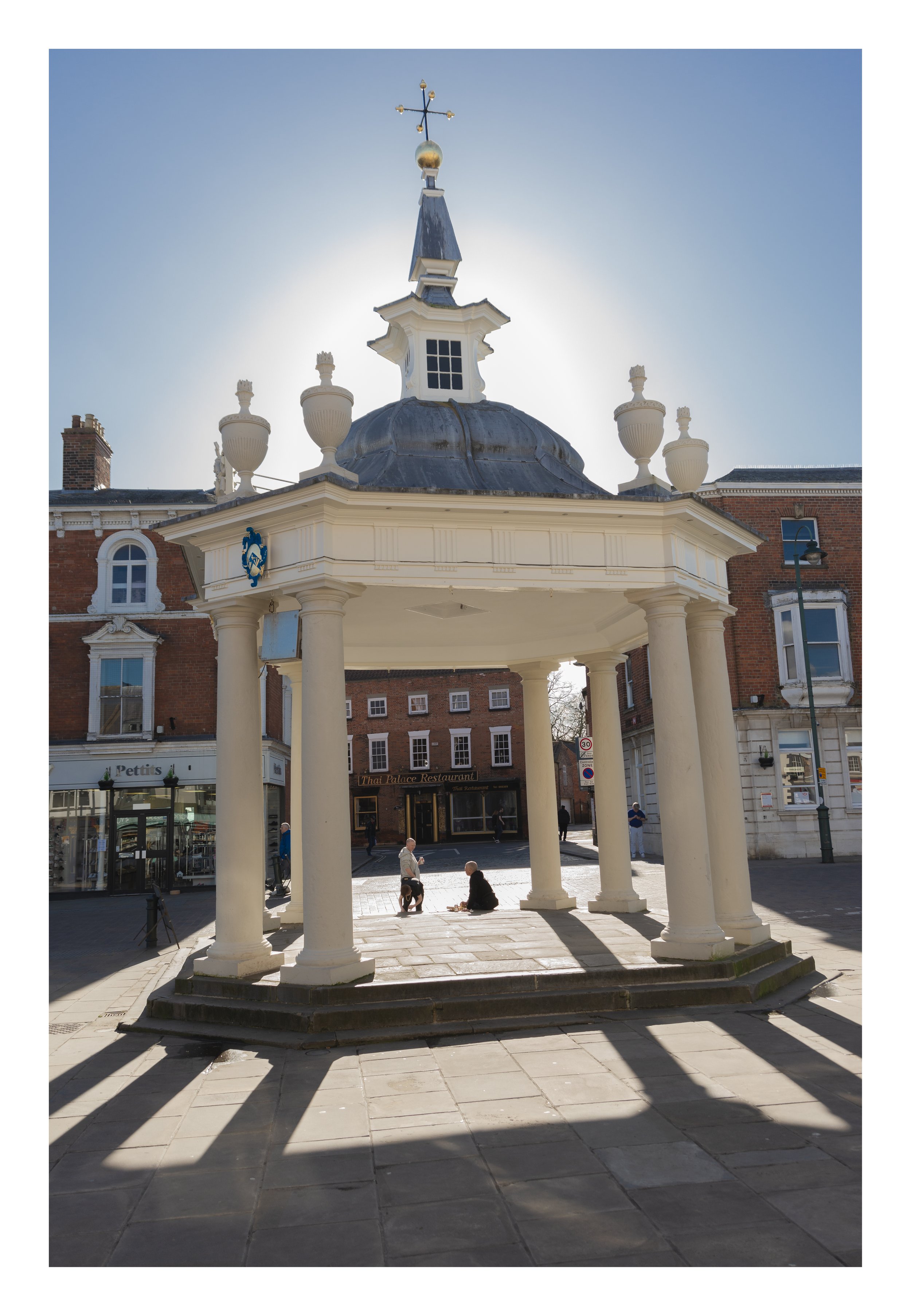 A white, classical-style pavilion with columns and a domed roof located in a town square. People are sitting and walking underneath it. In the background, there are brick buildings with shops and windows, a streetlamp, and a clear blue sky.