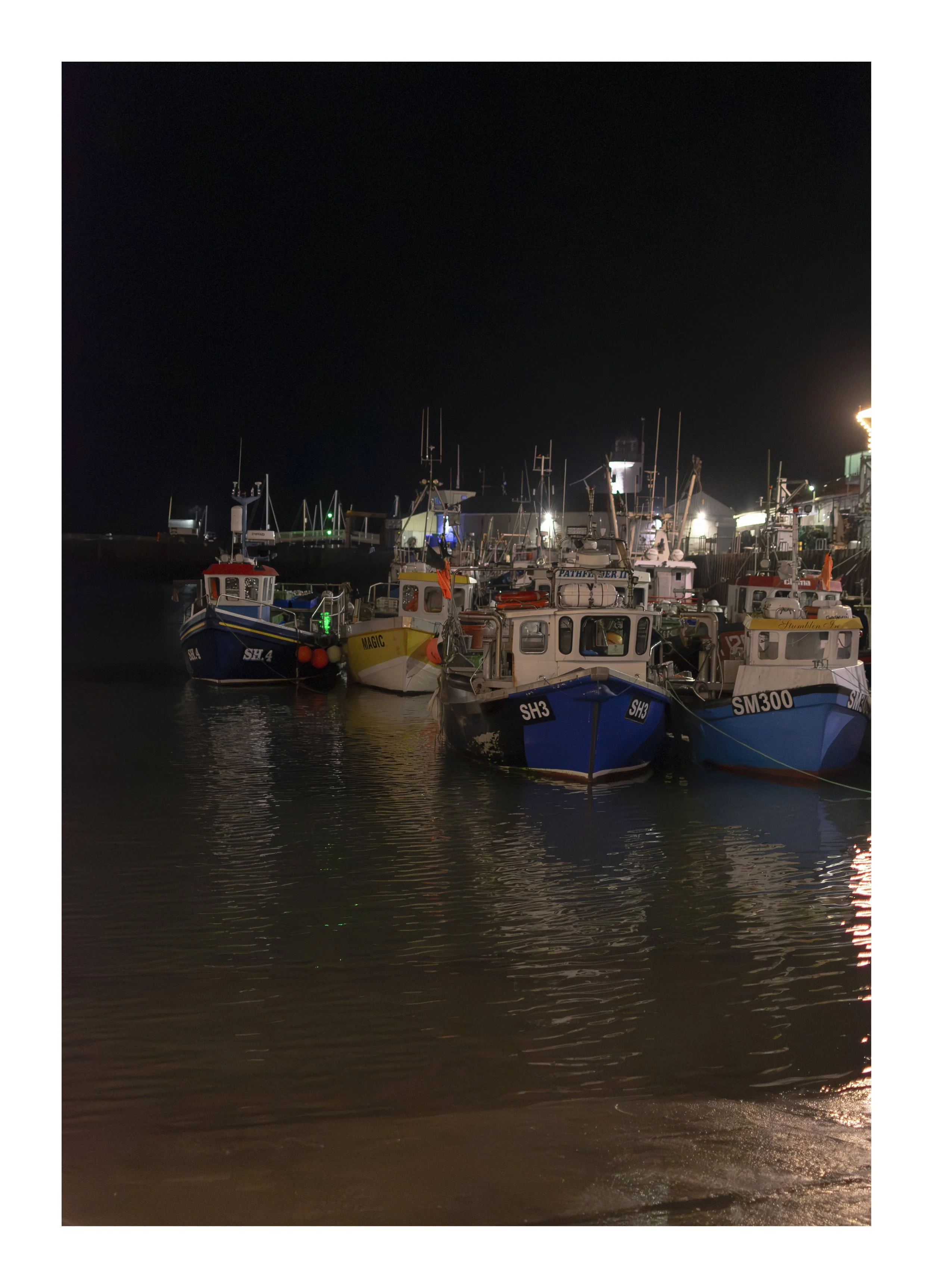 Nighttime view of boats docked at a marina with calm water and reflections, under a dark sky.
