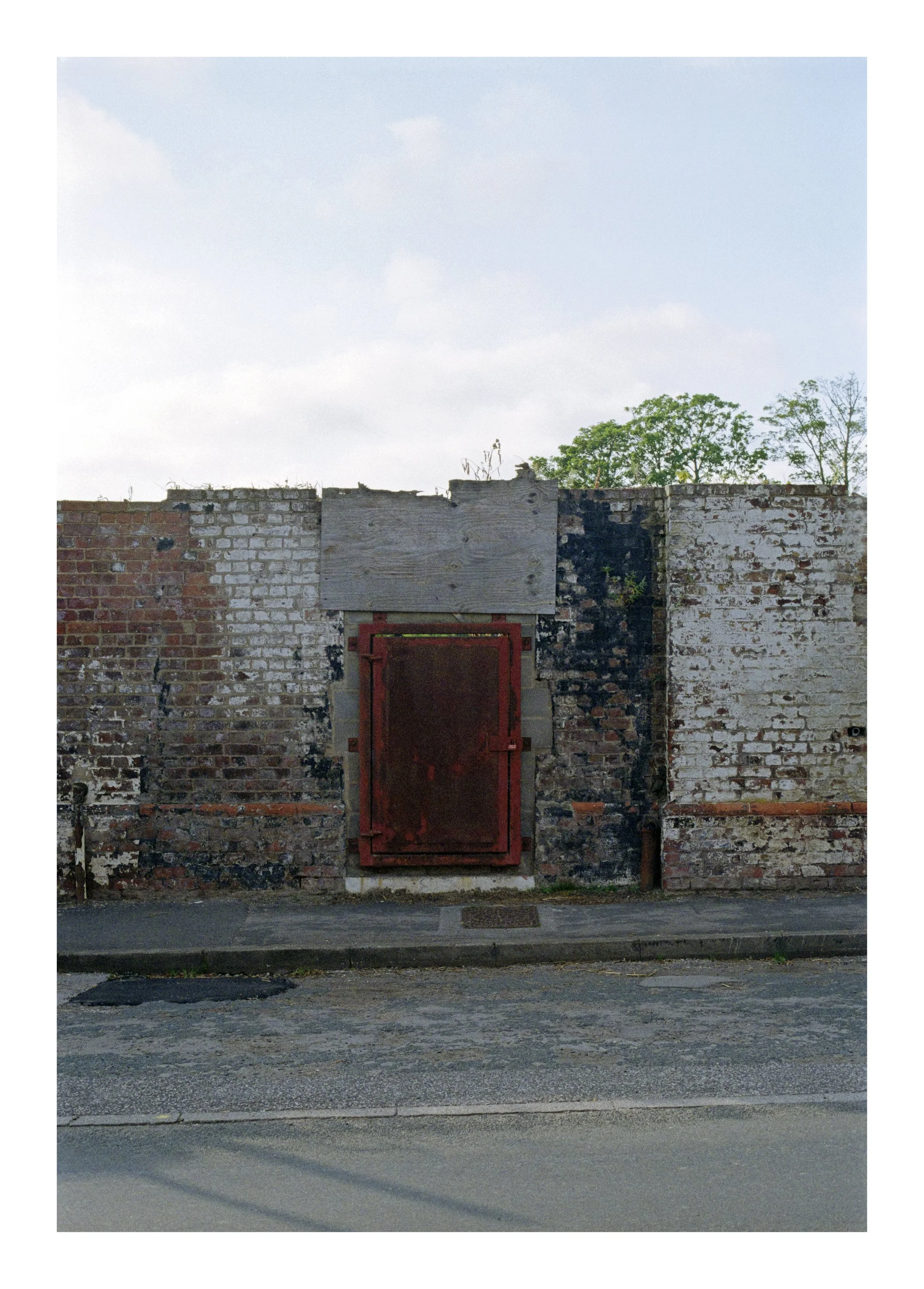 A brick wall with a boarded-up section and a red metal door, located beside a sidewalk with a street and patches of asphalt in the foreground. Trees and a cloudy sky are visible in the background.