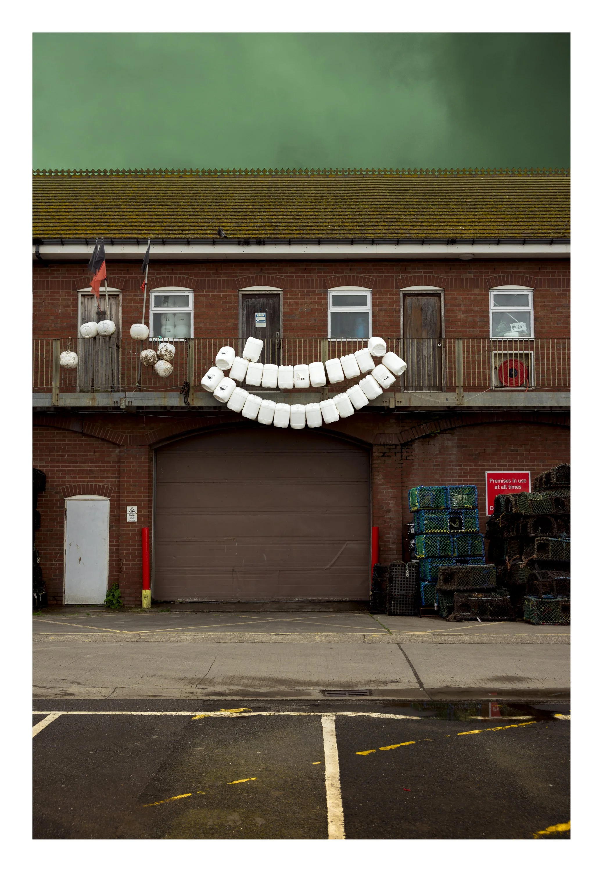 Red brick building with a large garage door, four small windows, and a balcony with white plastic containers arranged in a smiley face pattern. There are flags, a fire hose reel, stacked plastic crates, and a sign in the parking lot.