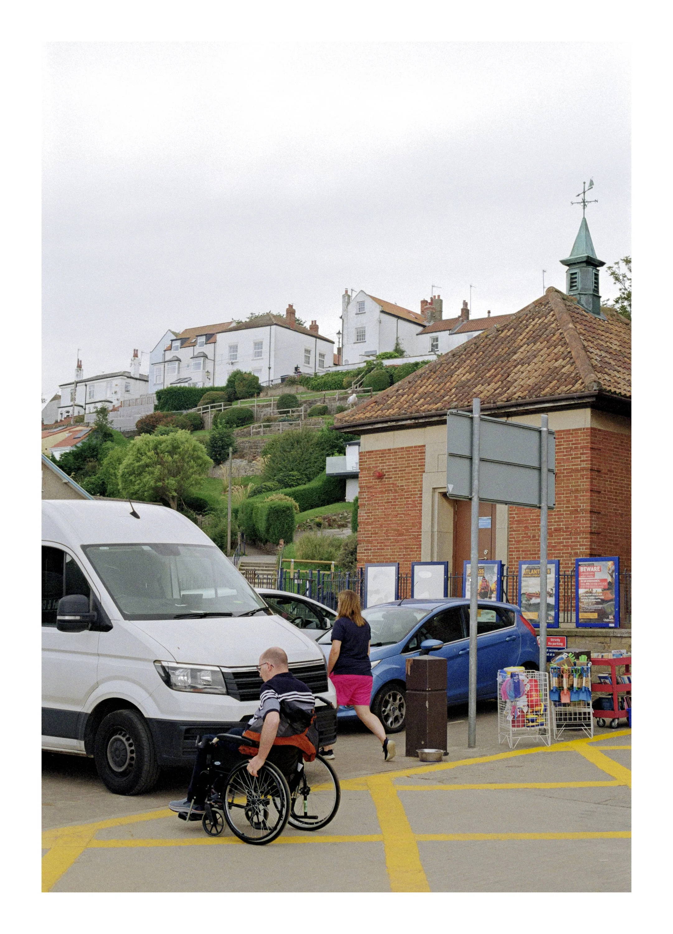 People in a parking lot, including a woman walking and a man in a wheelchair, surrounded by parked cars, a roadside stall with toys, and a building with a brick facade and a small clock tower or weather vane, with houses on a hill in the background.