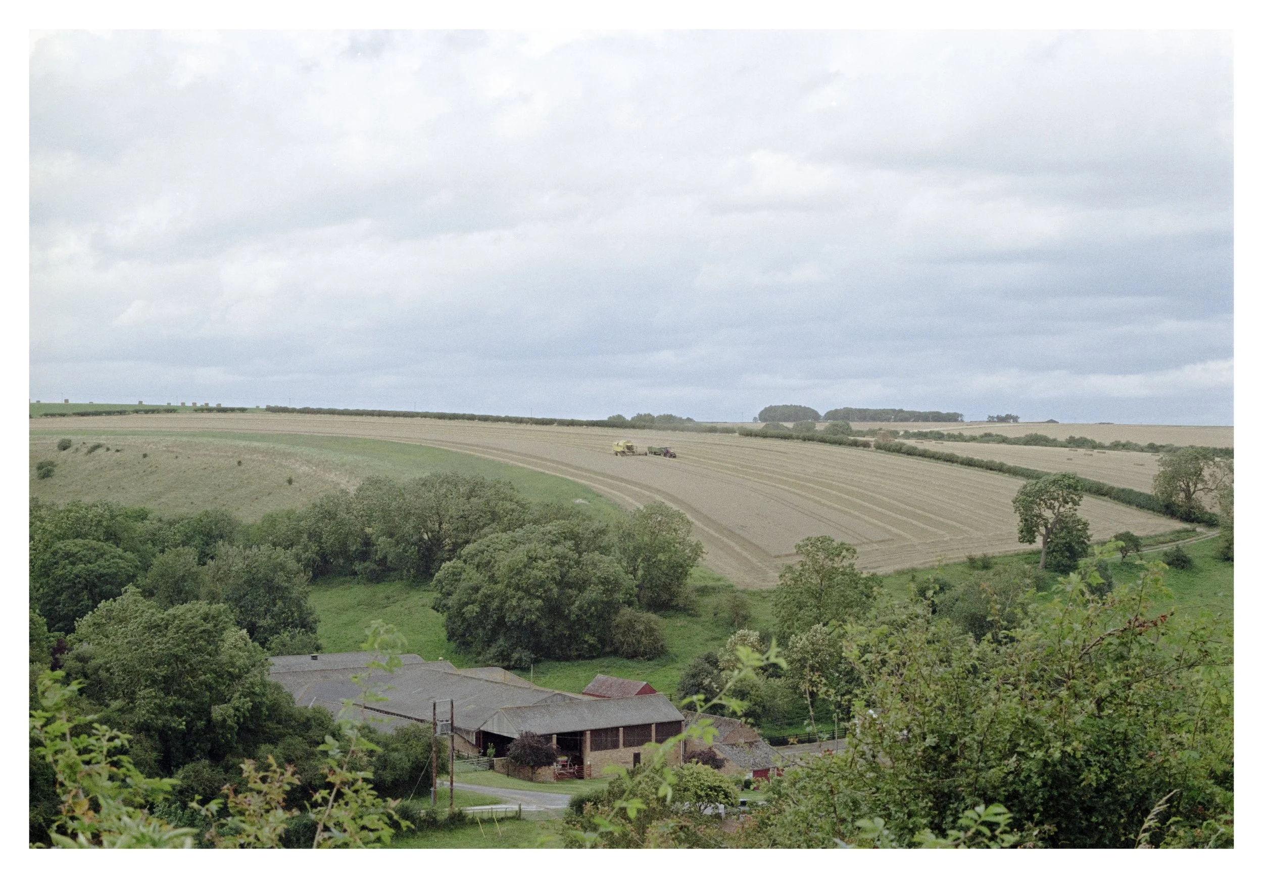 A rural landscape scene with a farm in the foreground and a wide open field in the distance under a cloudy sky.