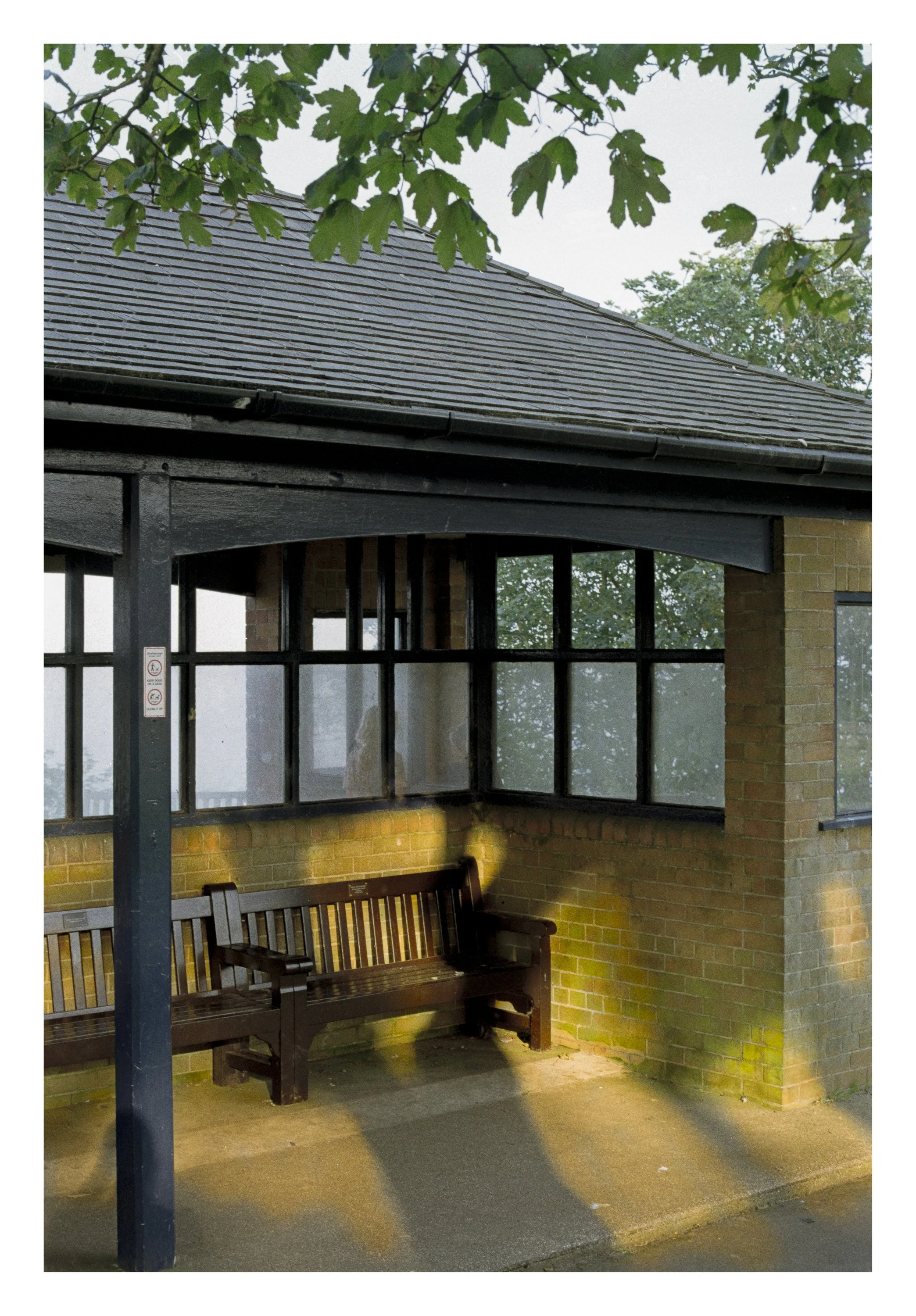 A covered outdoor seating area with a wooden bench, brick walls, large windows, and a sloped tiled roof, surrounded by greenery.