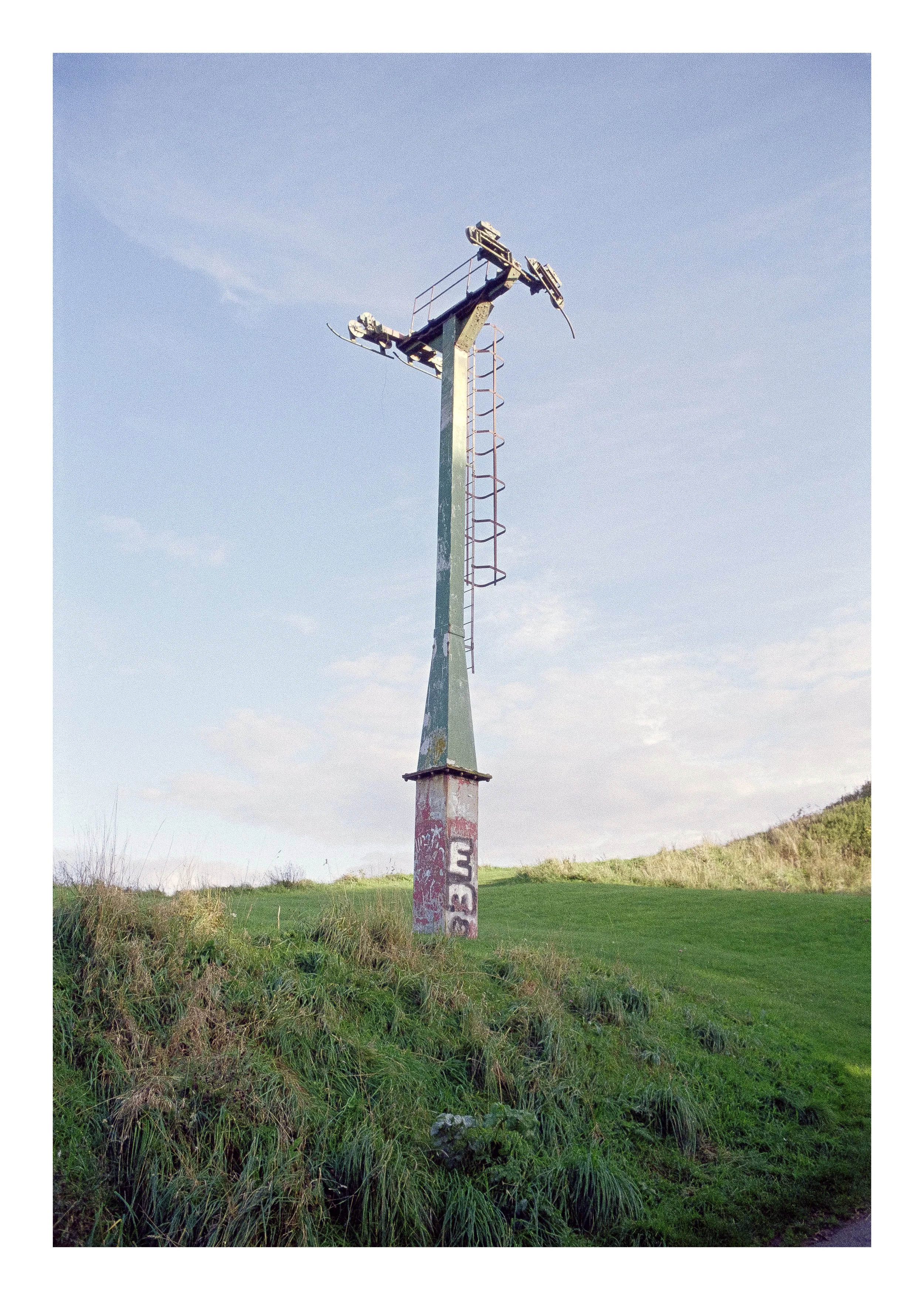 An old, weathered metal pole with decorative red honeycomb lattice ladders on its side, standing in a grassy field with a gentle hill in the background under a clear blue sky.