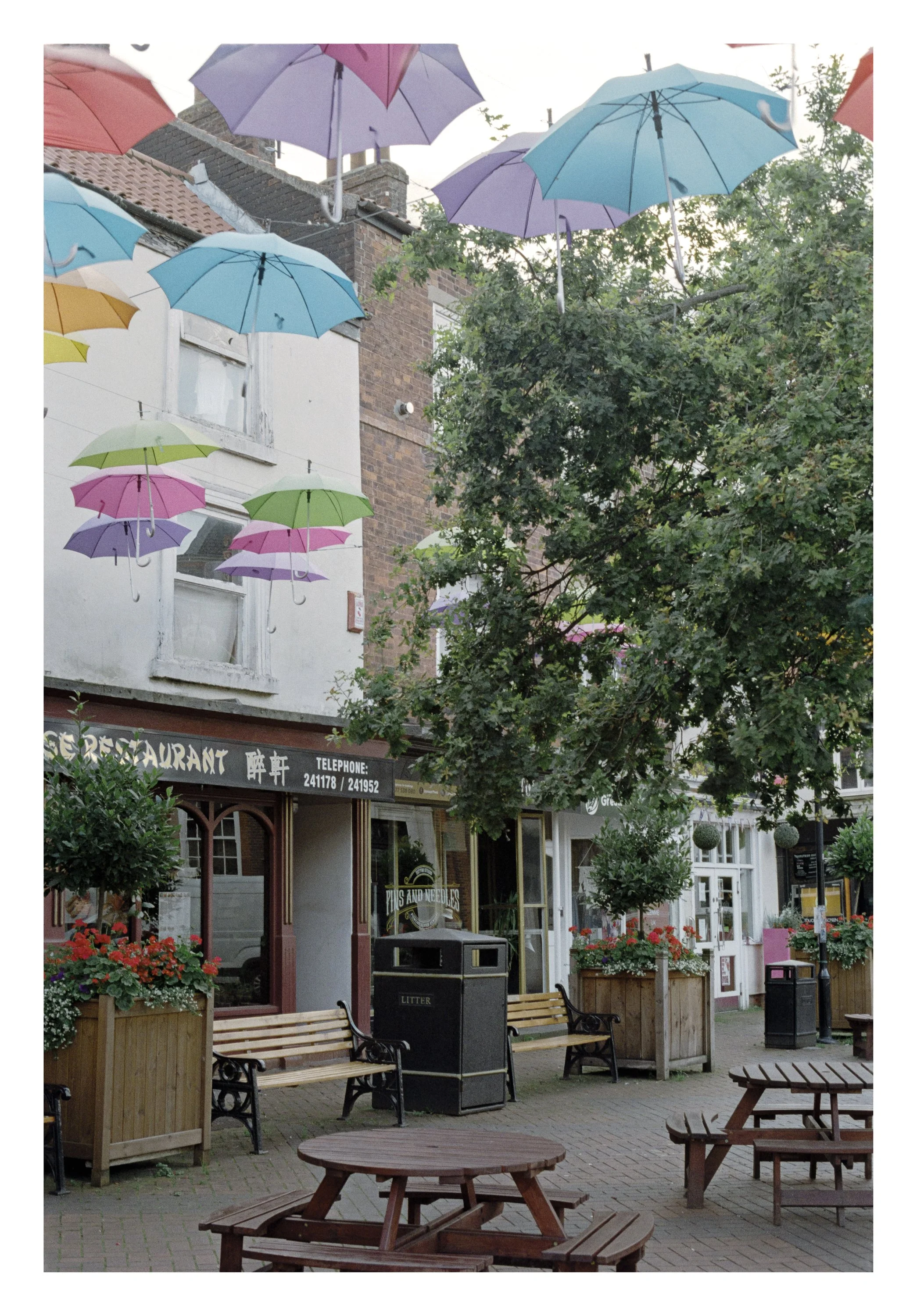 Outdoor sidewalk cafe with wooden tables and benches, decorated with colorful umbrellas hanging overhead, surrounded by plants and storefronts.