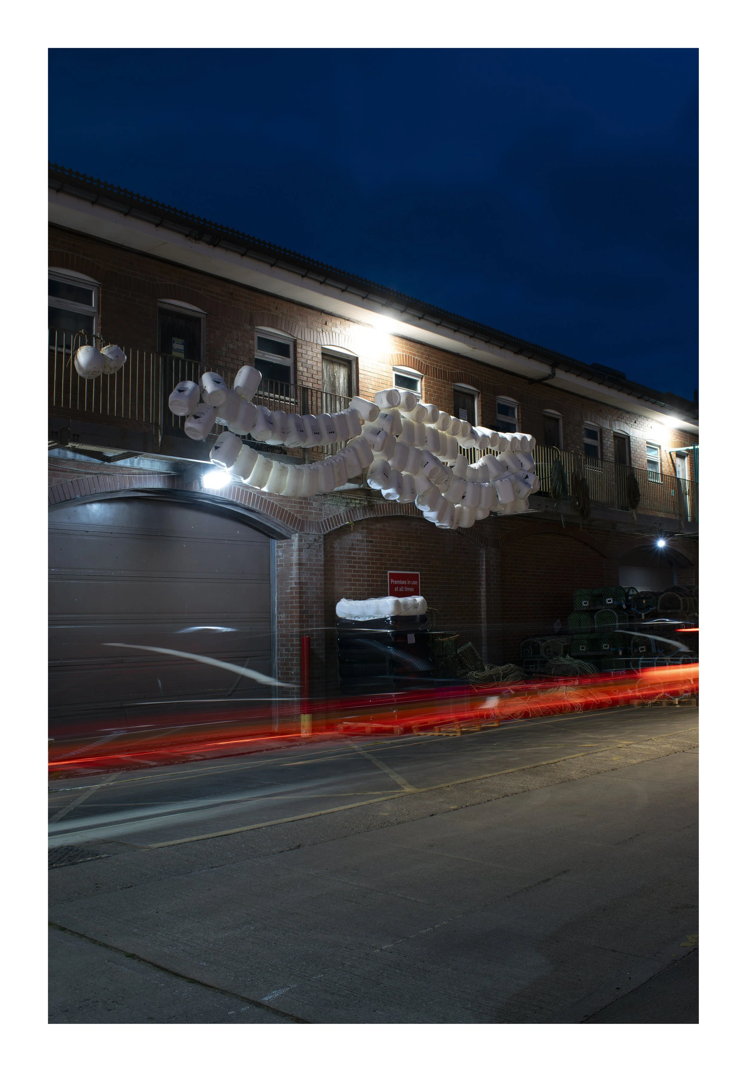 Nighttime street scene with a brick building featuring multiple windows and small balconies, decorated with a large string of white lanterns. The sky is dark blue, and the street has some motion blur from passing vehicles, with visible light streaks.