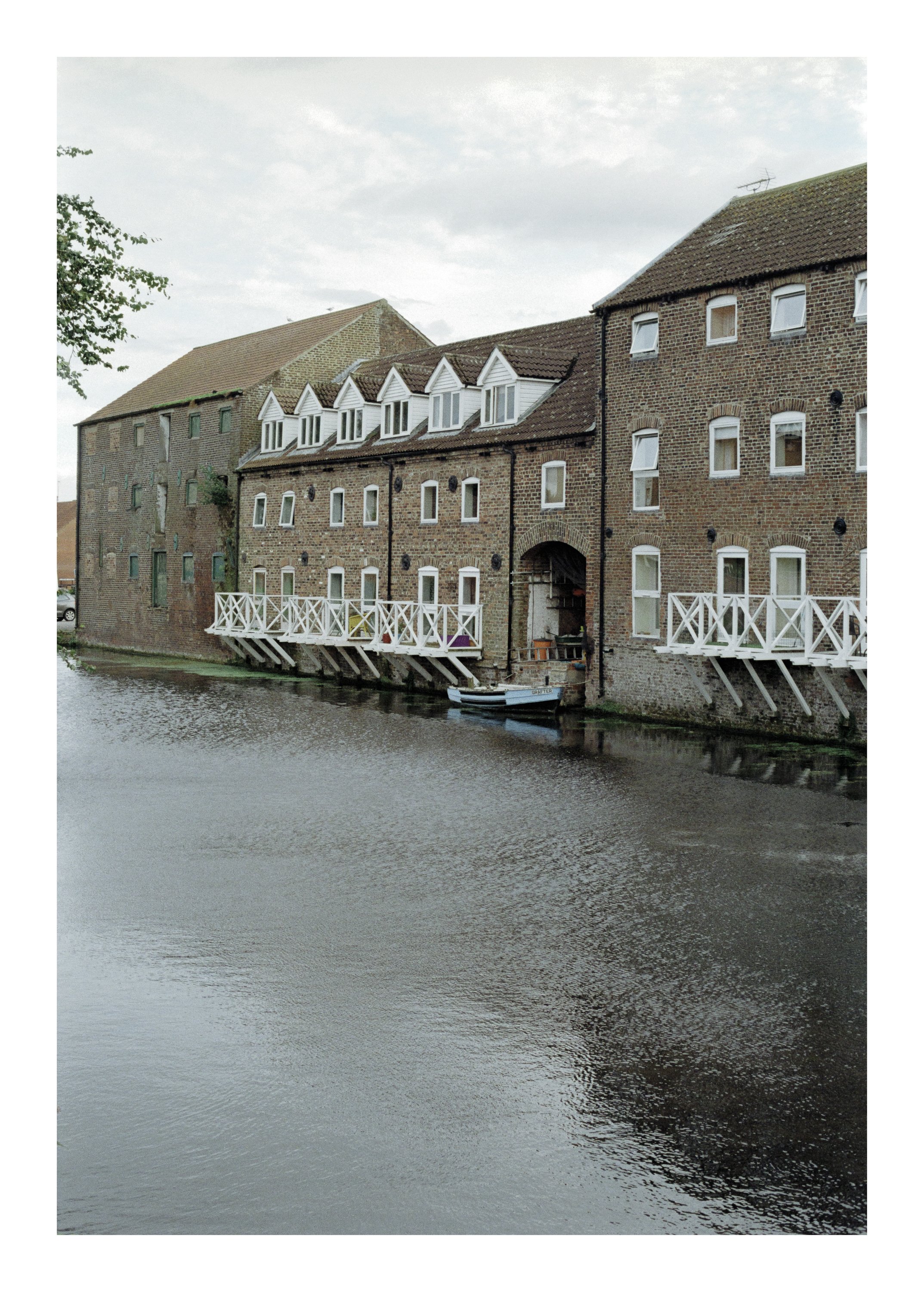 Brick buildings with white-framed windows and balconies along a river under a cloudy sky.