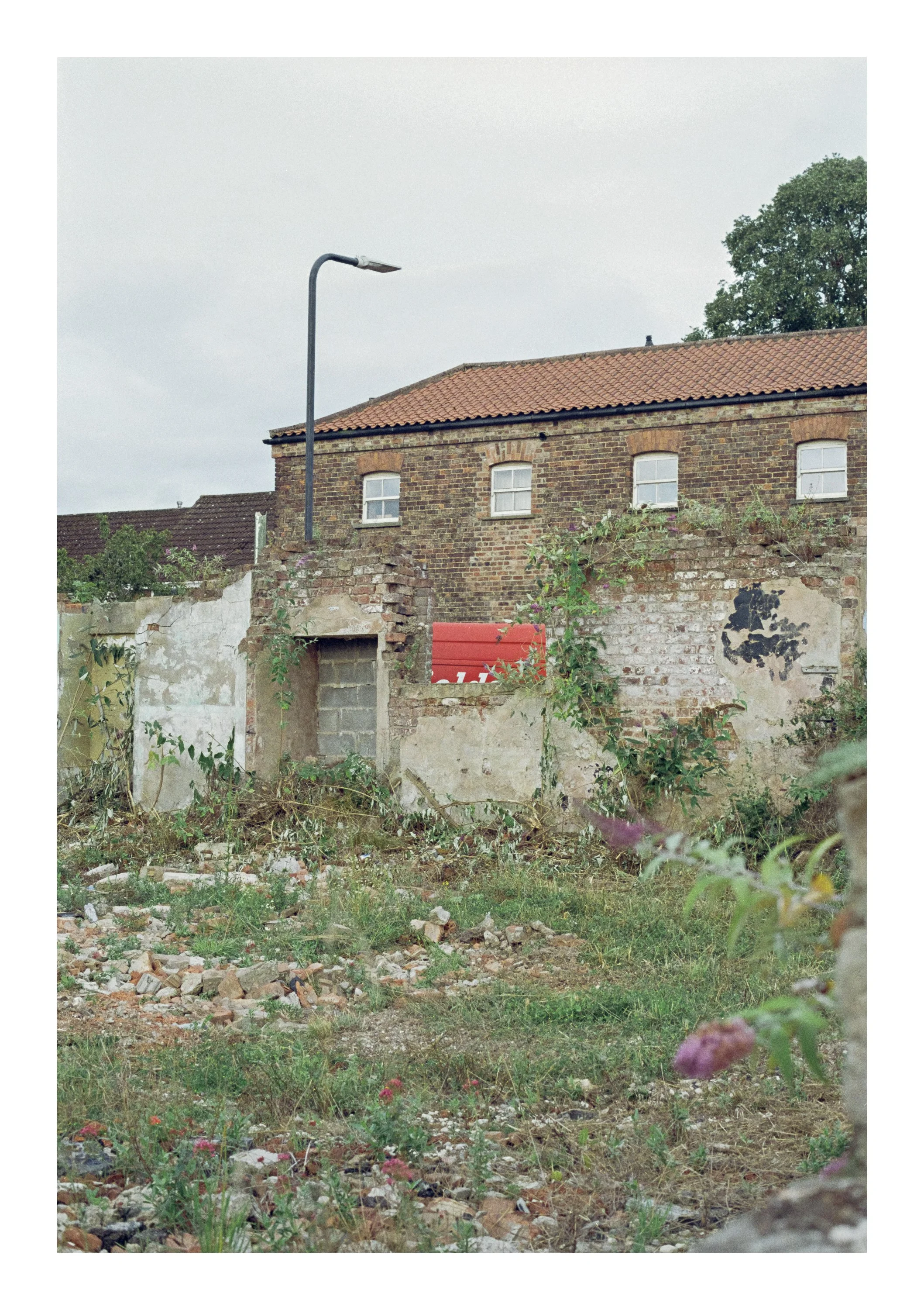 An old brick building with a red-tiled roof and small white windows, a street lamp, overgrown plants, and a wall that is partly crumbling surrounded by weeds and debris.