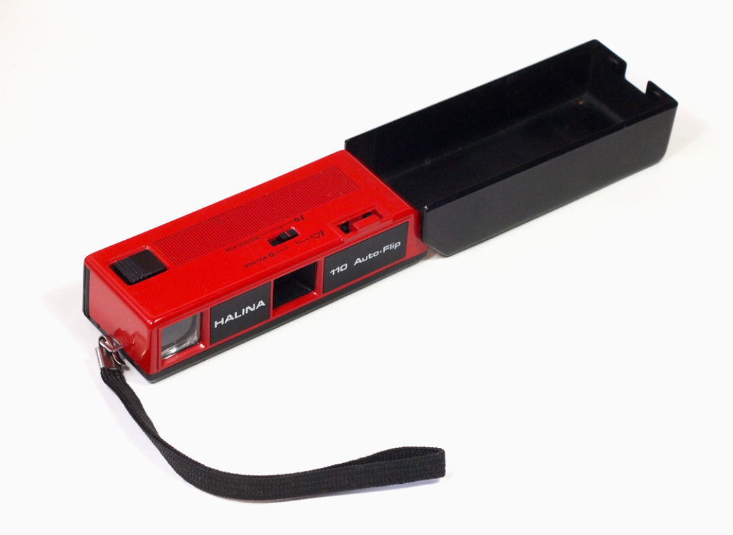 A vintage red and black Halina 110 Auto Flip 35mm film camera with a wrist strap, placed on a white surface.
