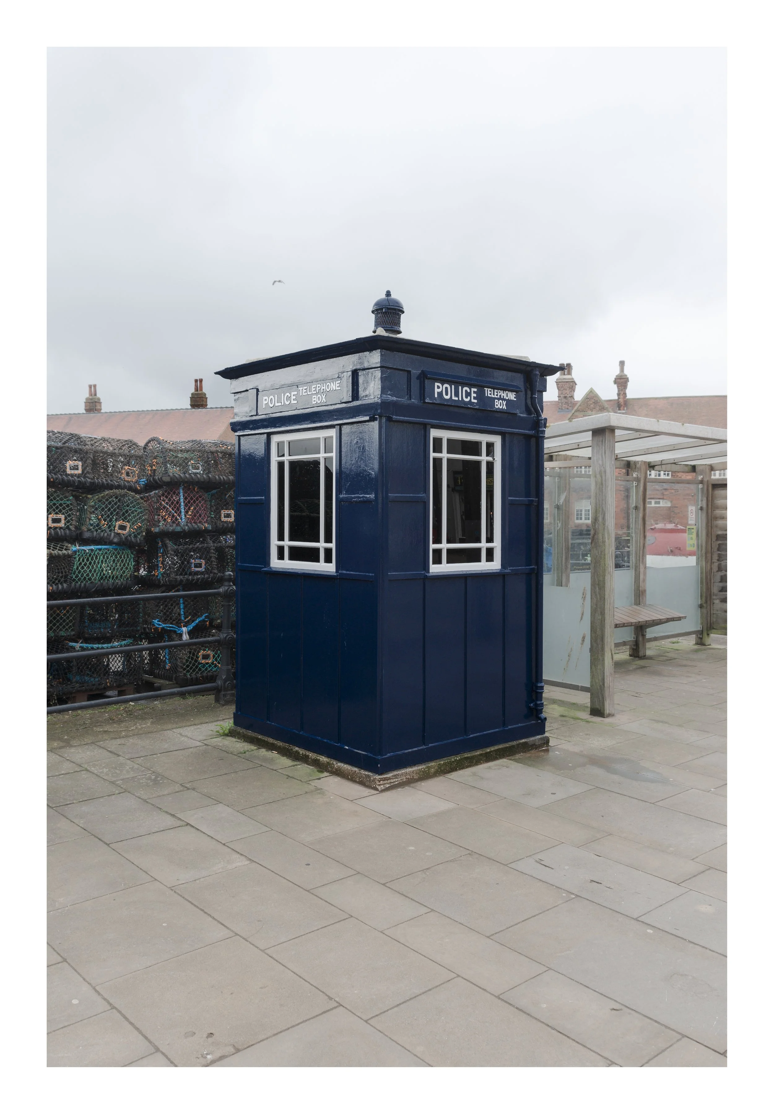A blue Police Telephone Box on a sidewalk, with a window on each side, surrounded by a brick-paved area and a background of buildings and cloudy sky.