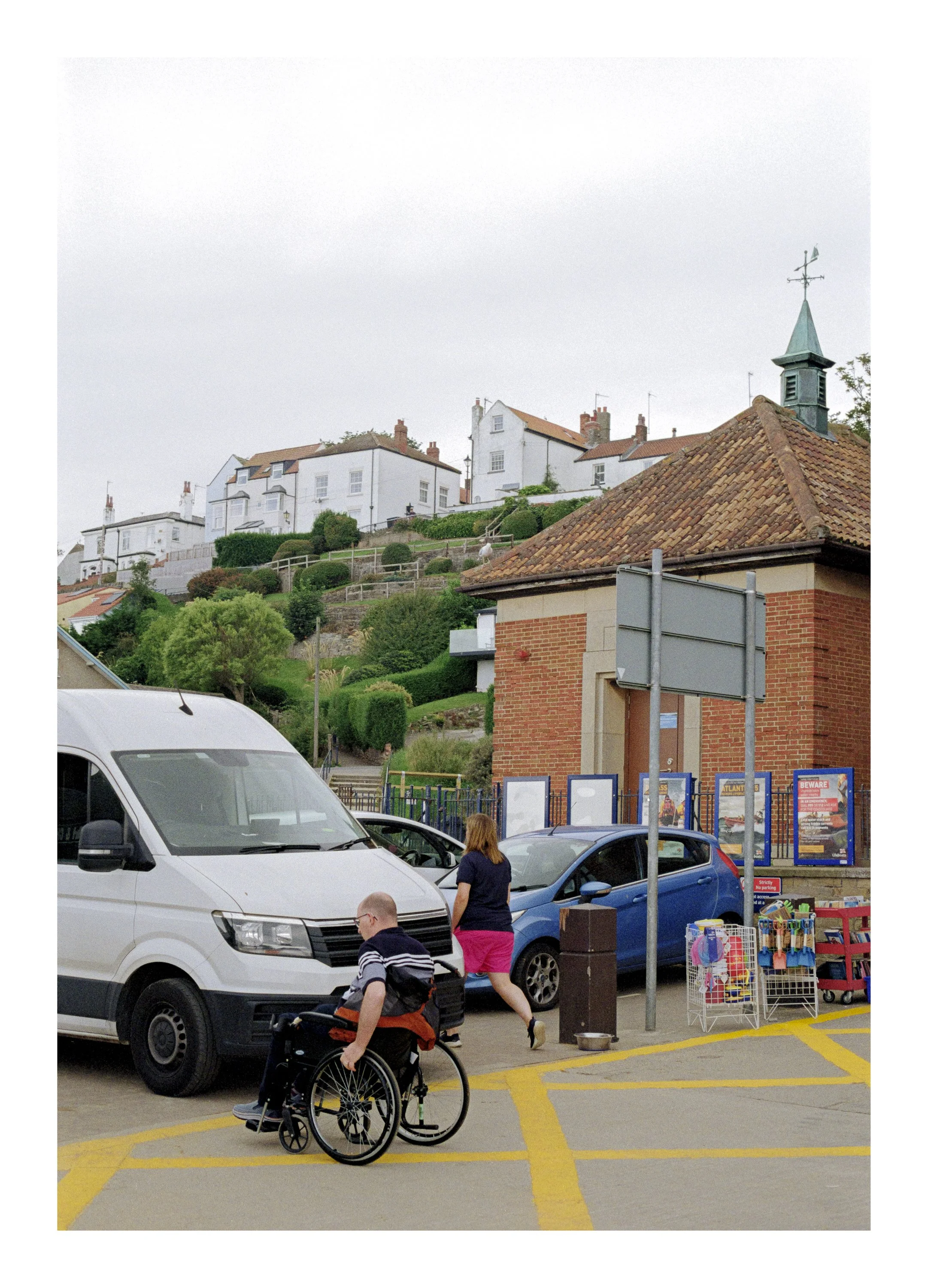 People walking and a person in a wheelchair in a parking lot near parked cars, a small street vendor stall with items for sale, and a hillside neighborhood with white houses and red roofs in the background.