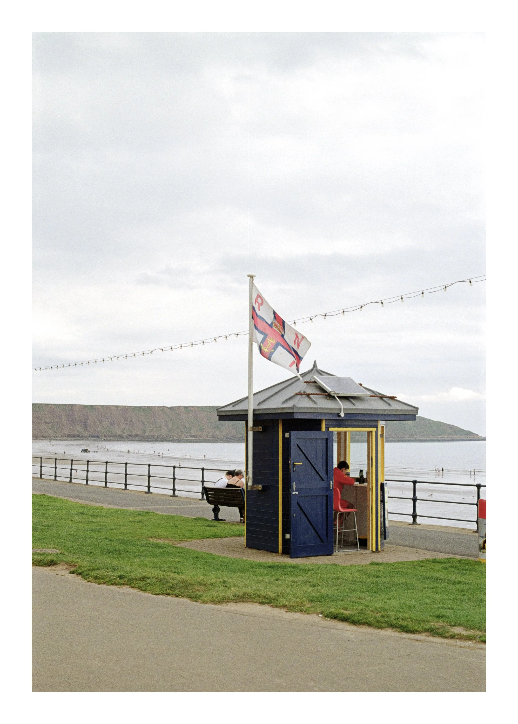 A small blue and yellow beach hut with an open door, situated along a walkway near the beach. There are two people seated on a bench, and another person in red clothing appears to be working inside the hut. In the background, the beach, ocean, and di