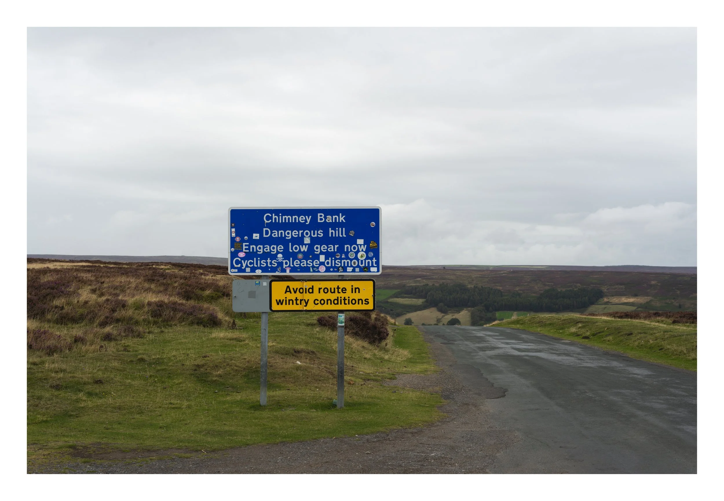 A blue road sign on a rural road with green fields in the background, warning about a chimney bank, dangerous hill, low gear, cyclists dismounting, and advising to avoid the route in wintry conditions.