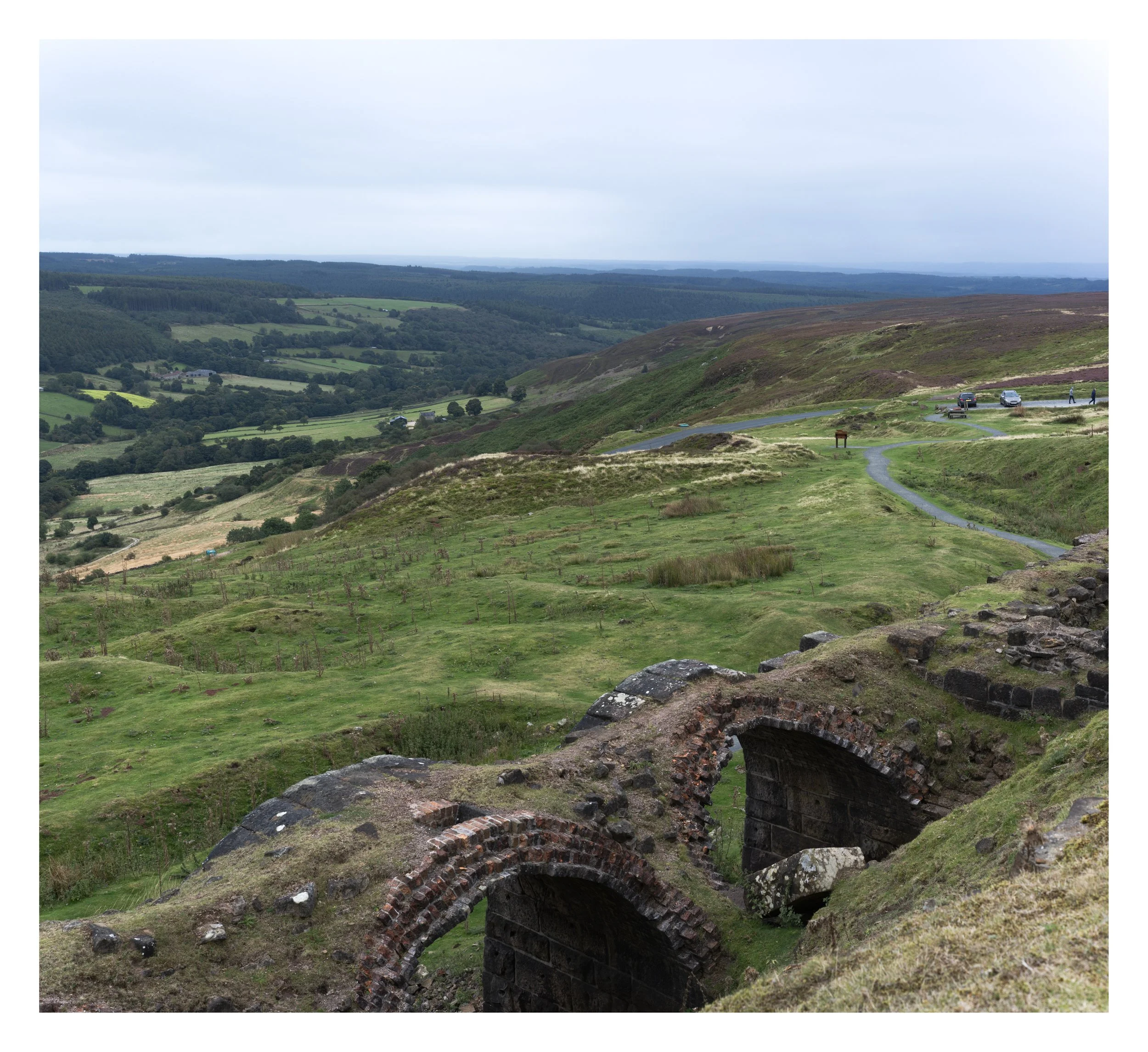 A landscape view of rolling green hills, forests, and a cloudy sky with some cars and a person walking on a path.