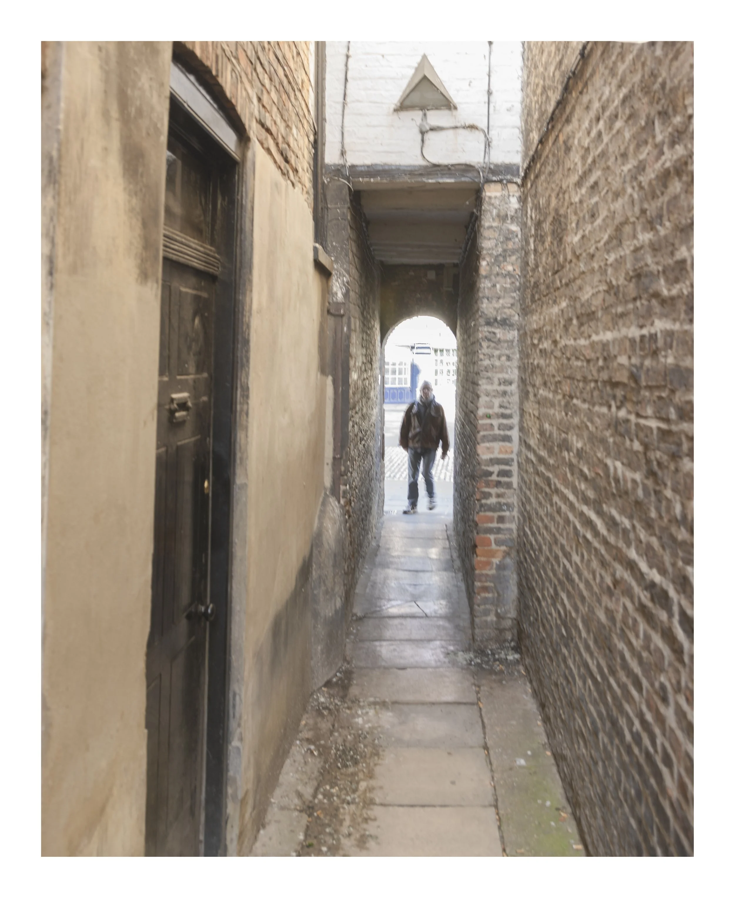 A narrow alleyway between brick and concrete buildings, with a person walking towards the camera in the background. The alley has a rough, dirty ground with some debris.