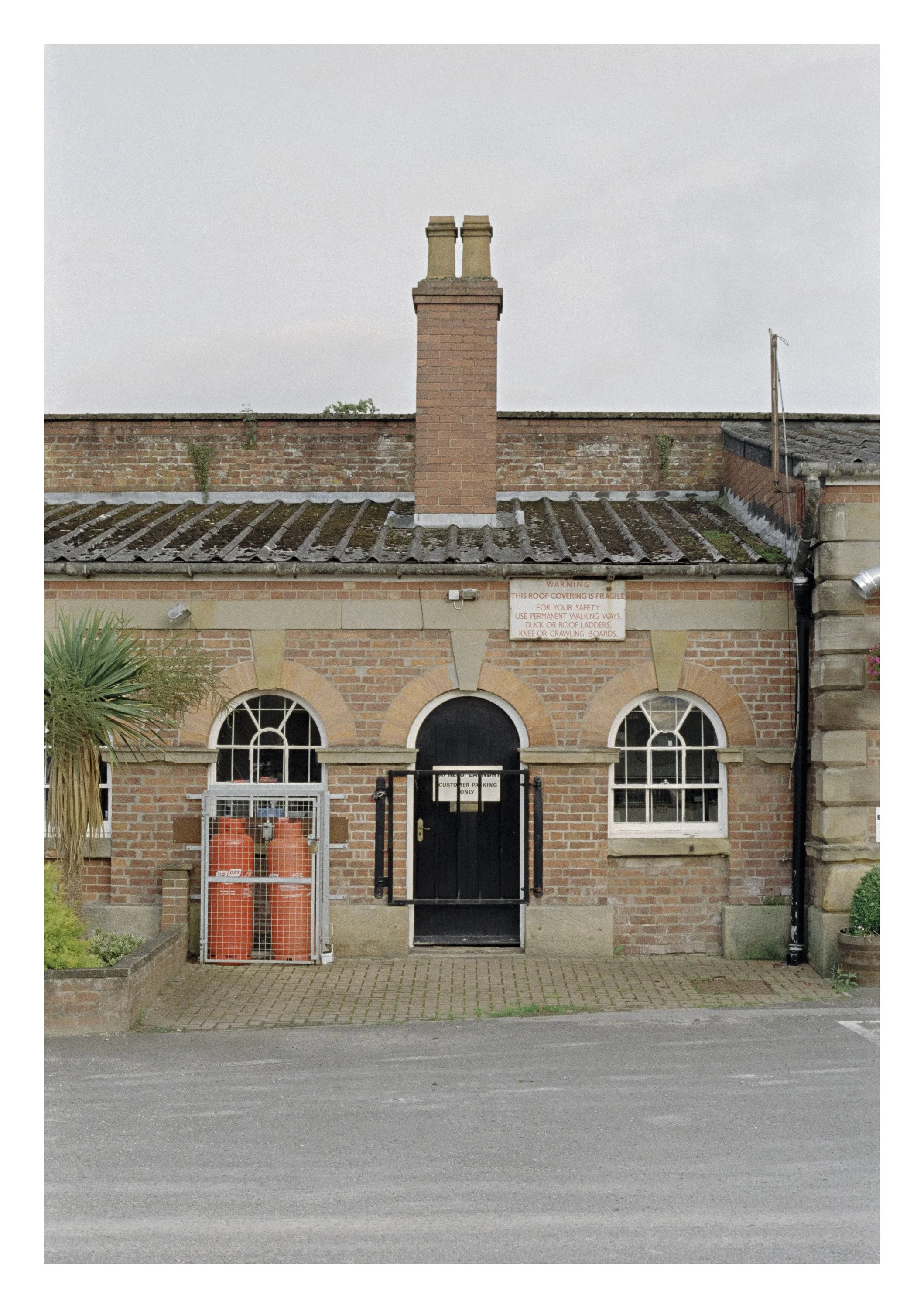 A brick building with a black gate, a sign about roof safety, a gas cage with two orange gas canisters, a plant on the left, and a paved driveway in front.
