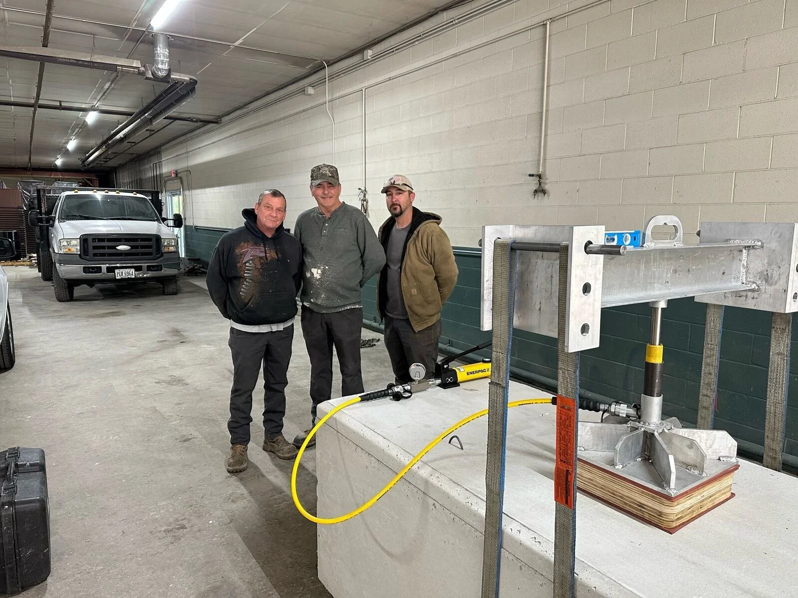 Three men standing inside a garage next to a large piece of equipment with a yellow hose, with trucks parked in the background.