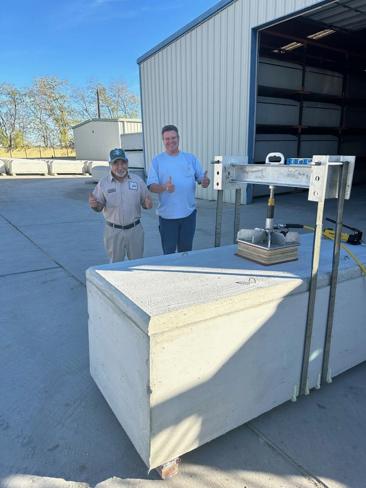 Two men standing outdoors next to a large stone block and a metal testing device, both giving thumbs up and smiling.