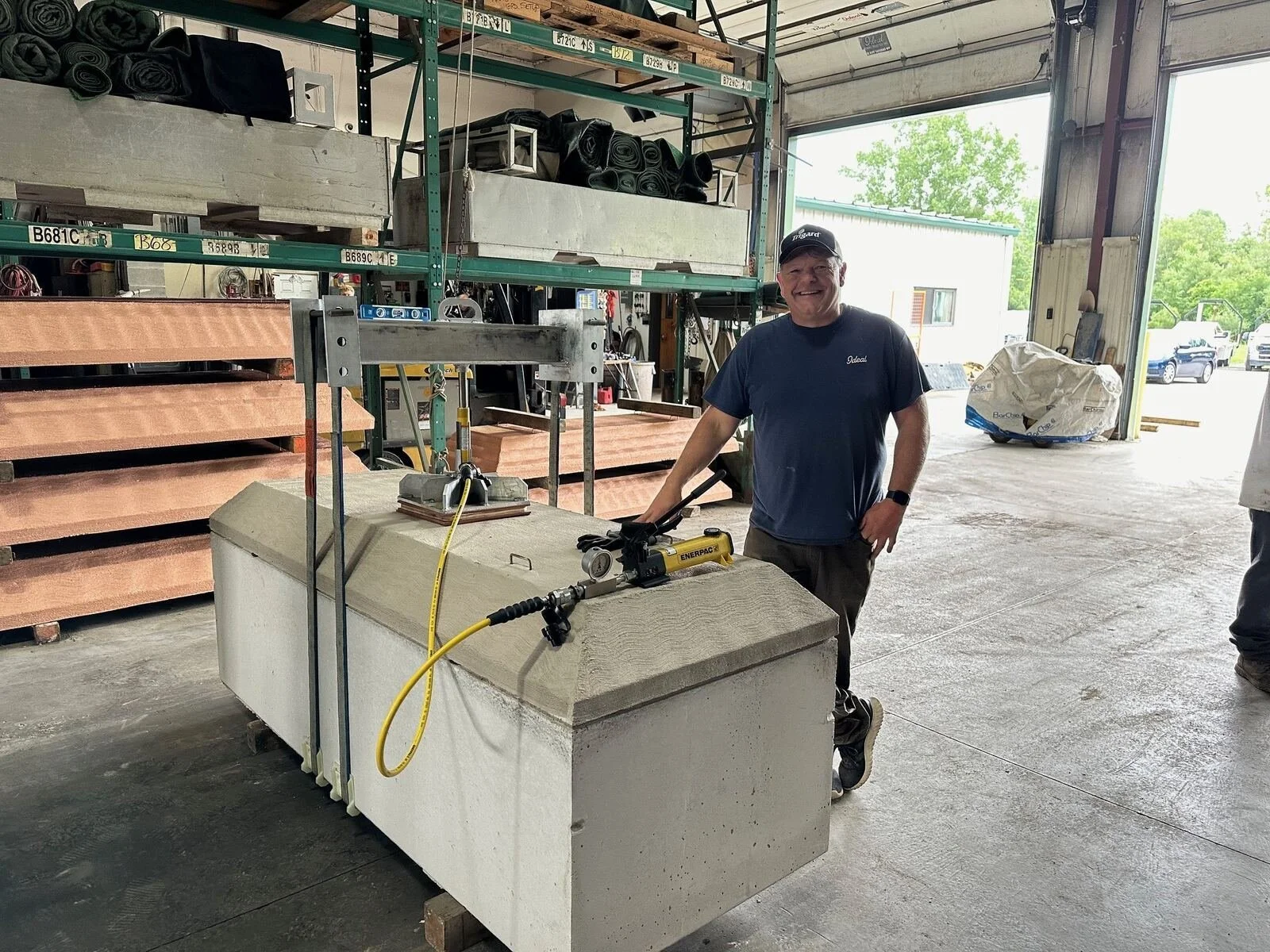 A man stands in a workshop with a large machine in front of him. The workshop has shelves filled with various materials and supplies. The man is smiling and wearing a cap and a dark shirt. There is an open door in the background with vehicles outside.